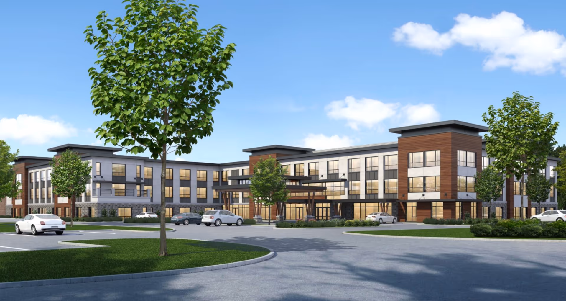 Modern three-story senior living facility building with large windows, wood and stone exterior accents, surrounded by green trees and a parking lot with several cars under a blue sky with scattered clouds.