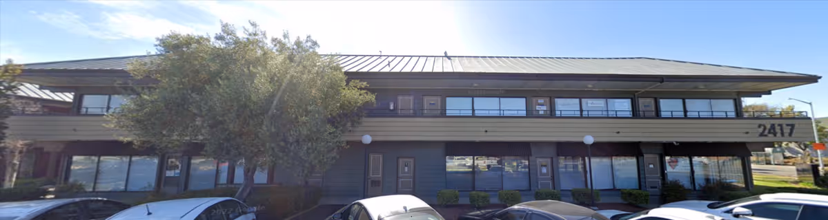 Two-story building with broad windows, a tree, and several parked cars in front.
