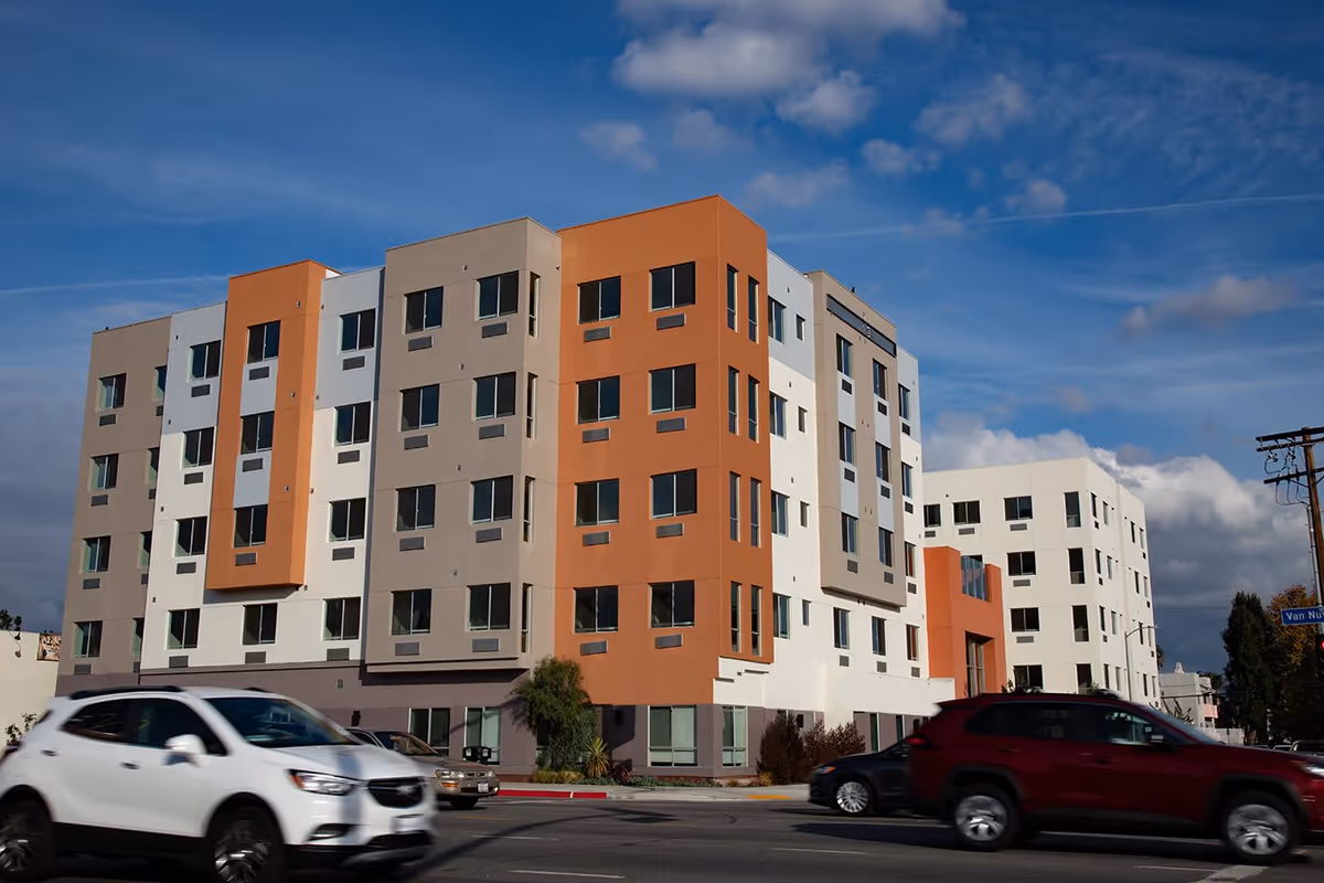 Street-level exterior view of a multi-story modern senior living building with orange and white facade and cars passing in front.