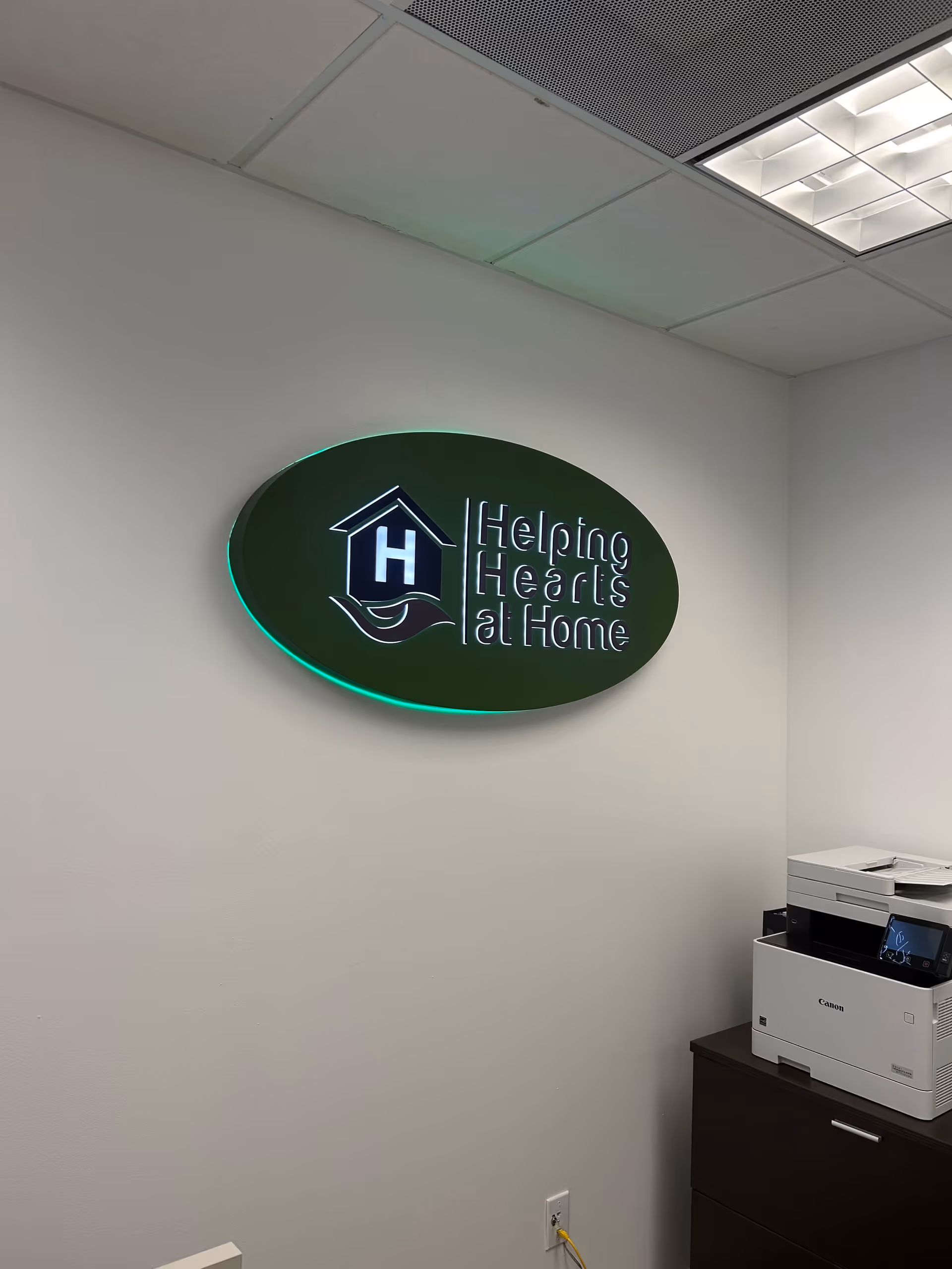 Interior hallway wall with illuminated Helping Hearts At Home sign and a printer on a cabinet.