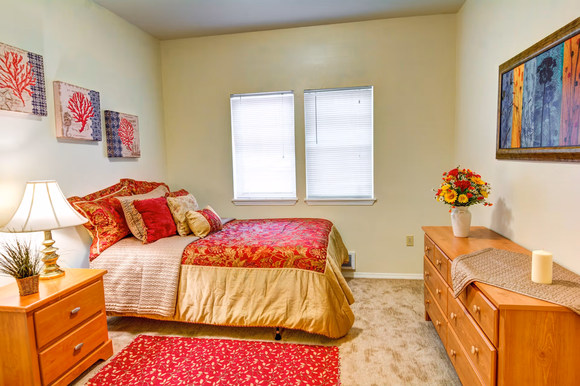 A cozy bedroom with a bed covered in red and gold bedding, multiple pillows, a wooden nightstand with a lamp and a small plant, a wooden dresser with a vase of flowers and a candle, two windows with blinds, and framed artwork on the walls.