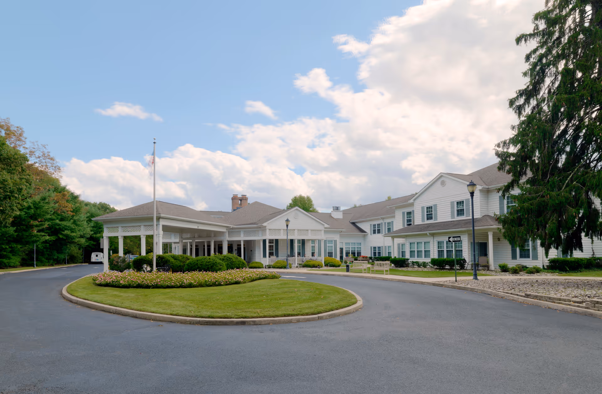 Exterior view of The Residence at Voorhees, a large white two-story building with a covered entrance, surrounded by a circular driveway and landscaped greenery under a partly cloudy sky.