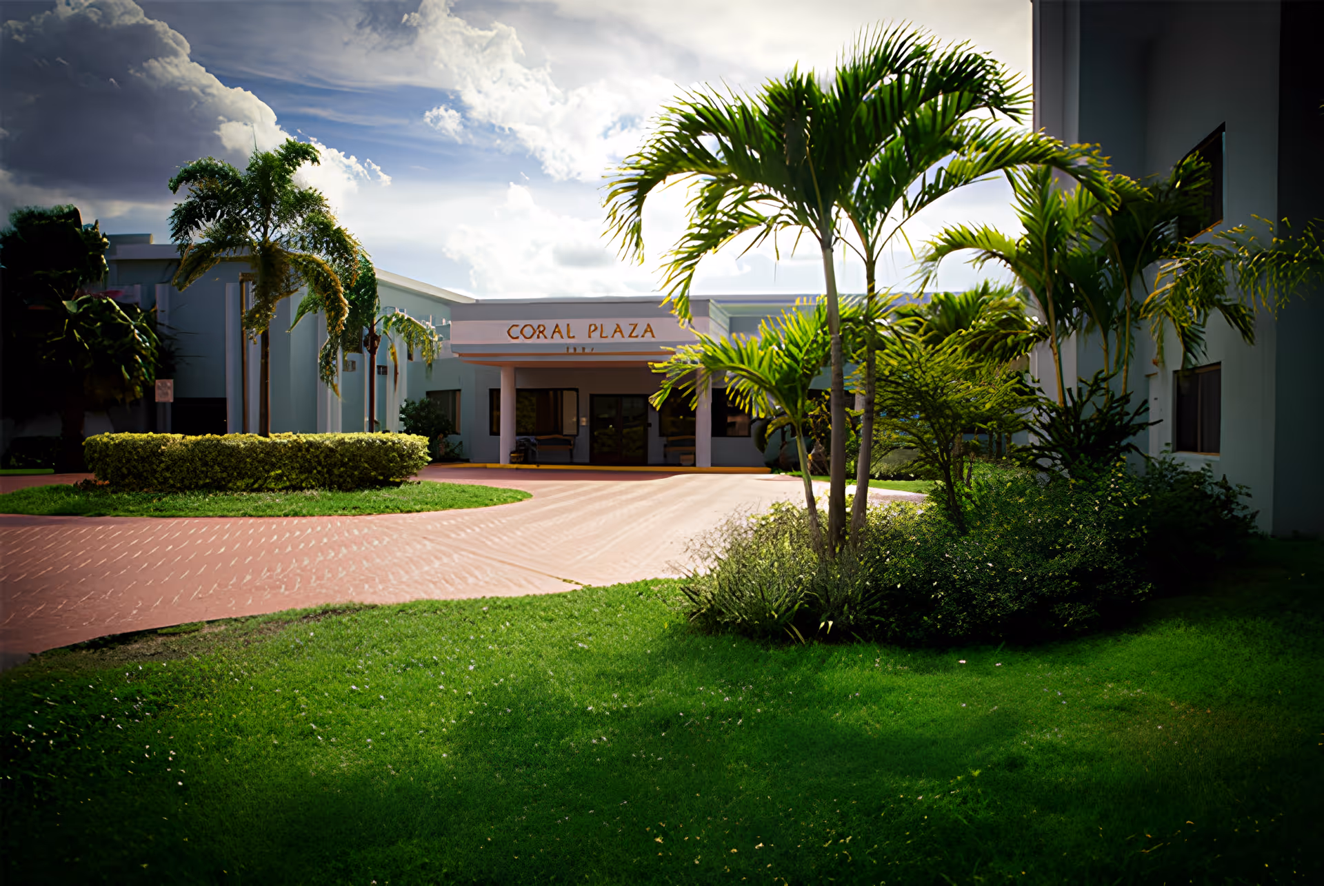 Entrance of Coral Plaza with a circular driveway, palm trees, and landscaped lawn in front of the building.