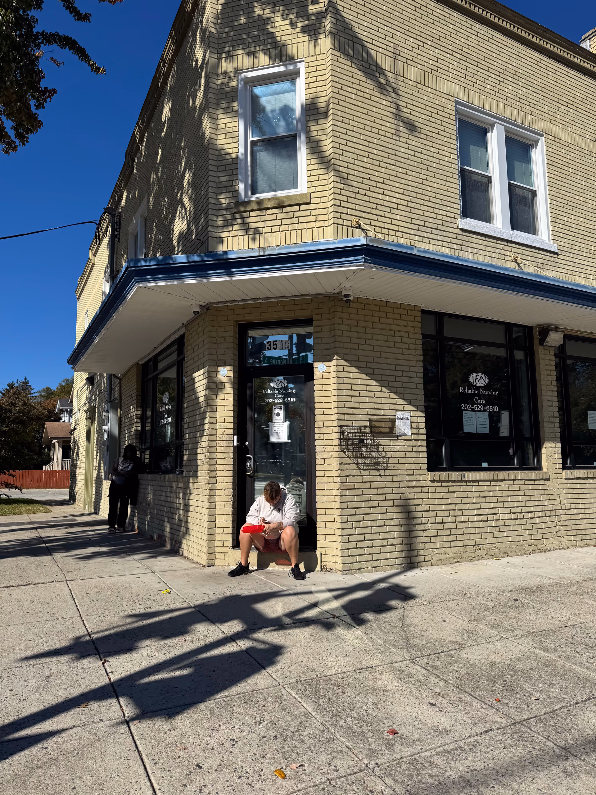 Two-story beige brick nursing care building with blue trim; a person sits on the front steps.