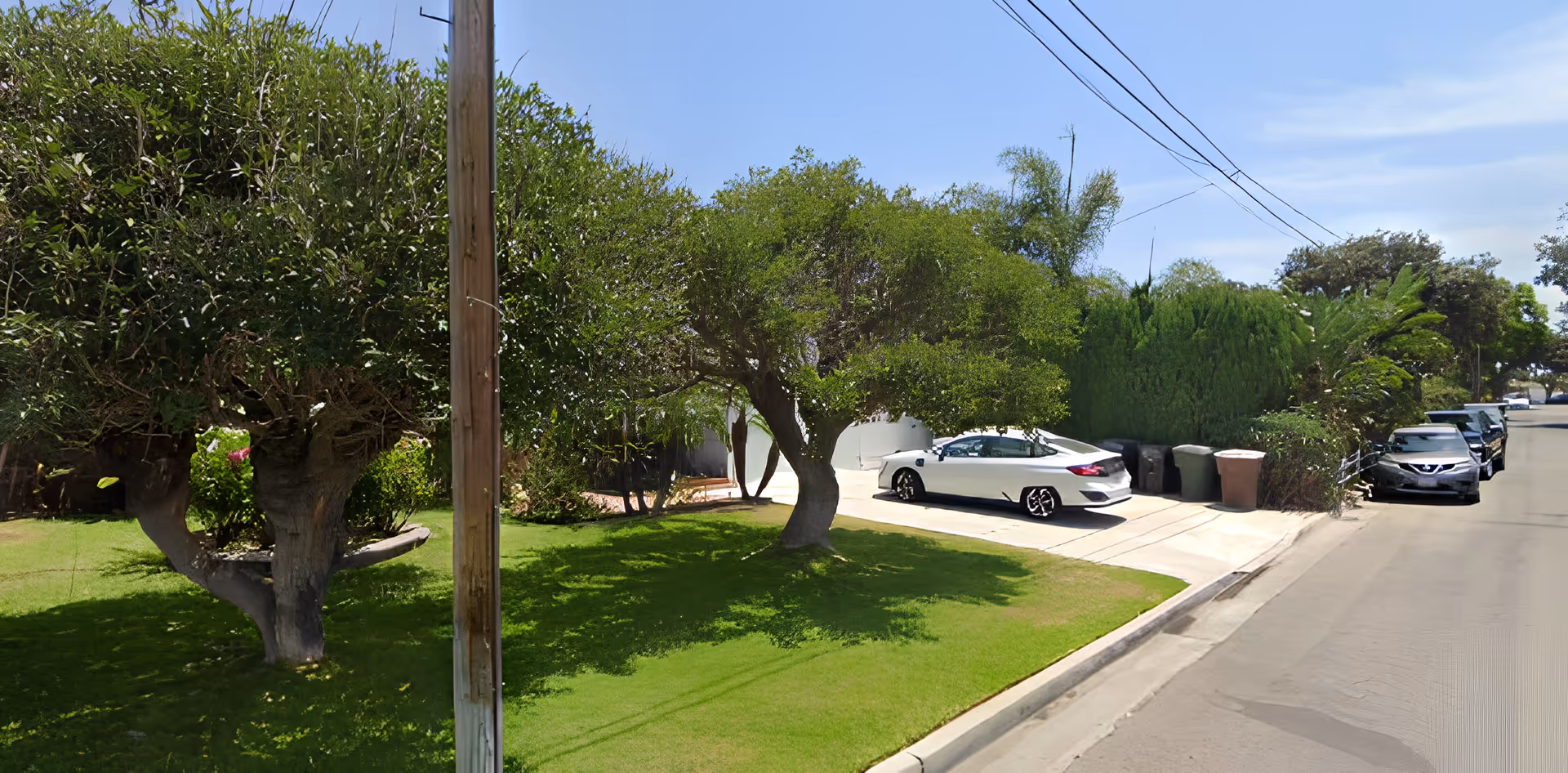 Street view of a house front with trimmed trees, a green lawn, a driveway holding a white car and other cars parked along the curb.