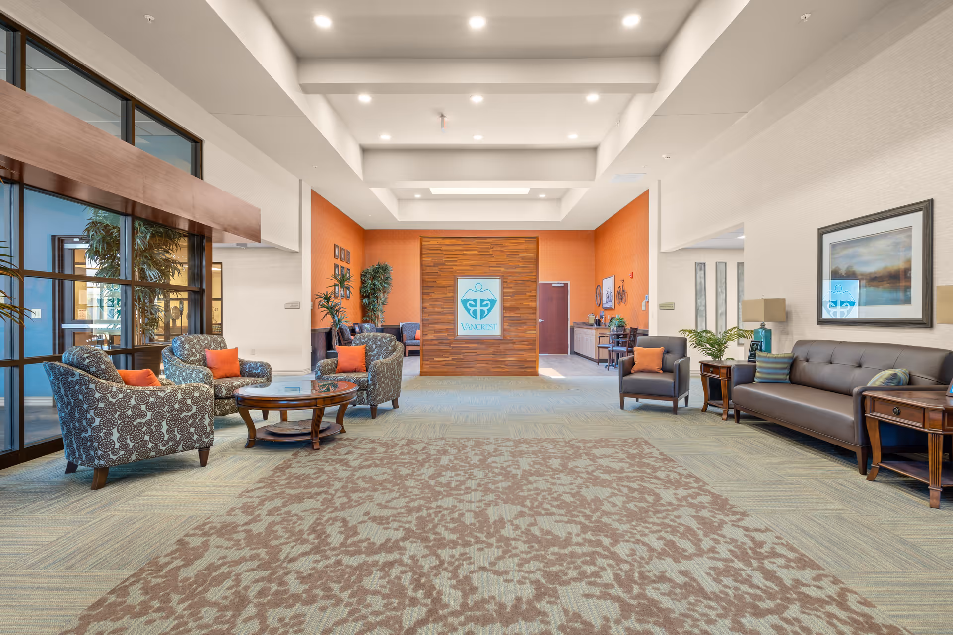 Spacious senior living lobby with seating area, plants, and a central wood-paneled wall logo.