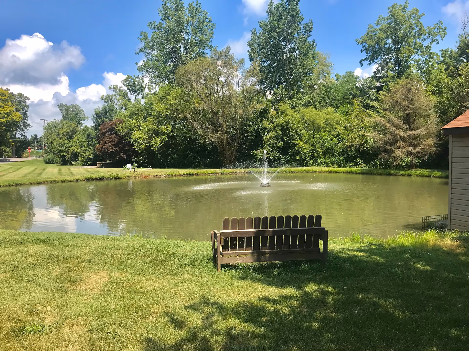 A peaceful outdoor scene featuring a small pond with a water fountain in the center, surrounded by green grass and trees. A wooden bench faces the pond, and a small shed is visible on the right side. The sky is blue with some clouds.