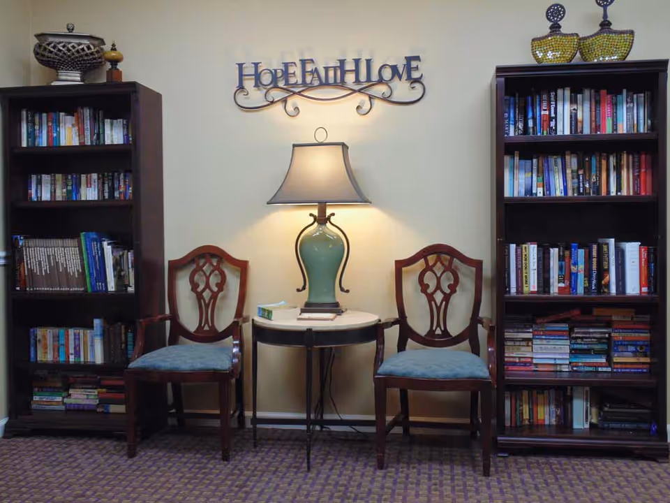 A cozy reading nook with two wooden chairs with blue cushions placed on either side of a small round table with a green lamp. Behind the table on the wall is a decorative sign that reads 'Hope Faith Love'. On each side of the chairs are tall dark wooden bookshelves filled with books and decorative items on top.