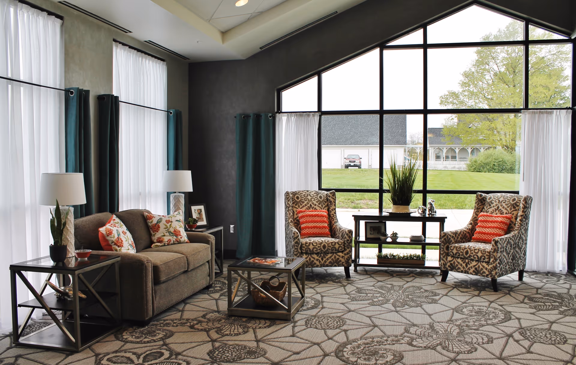 A cozy living room area with a brown sofa adorned with floral and red pillows, two patterned armchairs with red pillows, a glass coffee table, side tables with lamps and plants, and large windows with white and dark green curtains letting in natural light and showing a green lawn outside.