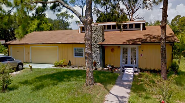 Single-story yellow building with a garage, stone chimney, ramped front entrance, and a grassy yard with trees.
