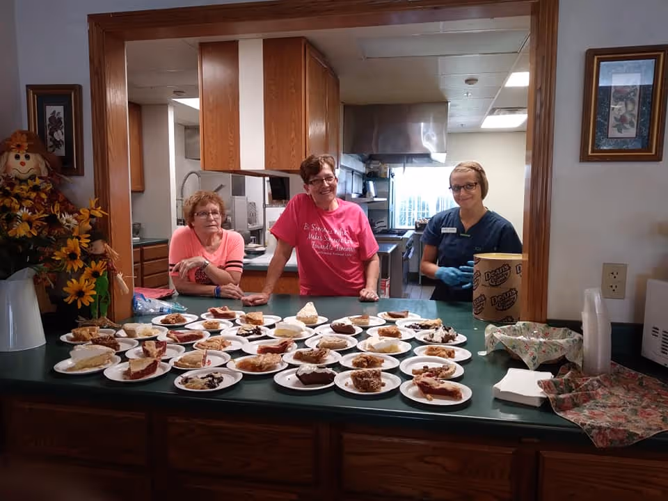 Three women standing behind a counter filled with various slices of pie on paper plates in a kitchen area. The woman on the left is wearing a pink shirt and glasses, the woman in the middle is wearing a bright pink shirt and glasses, and the woman on the right is wearing a dark blue uniform with gloves. The kitchen has wooden cabinets and stainless steel appliances. There are decorative flowers and framed pictures on the walls.