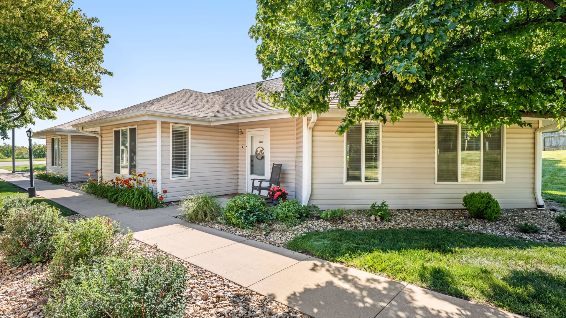 Single-story beige facility with front entrance, windows, and a landscaped path under trees.