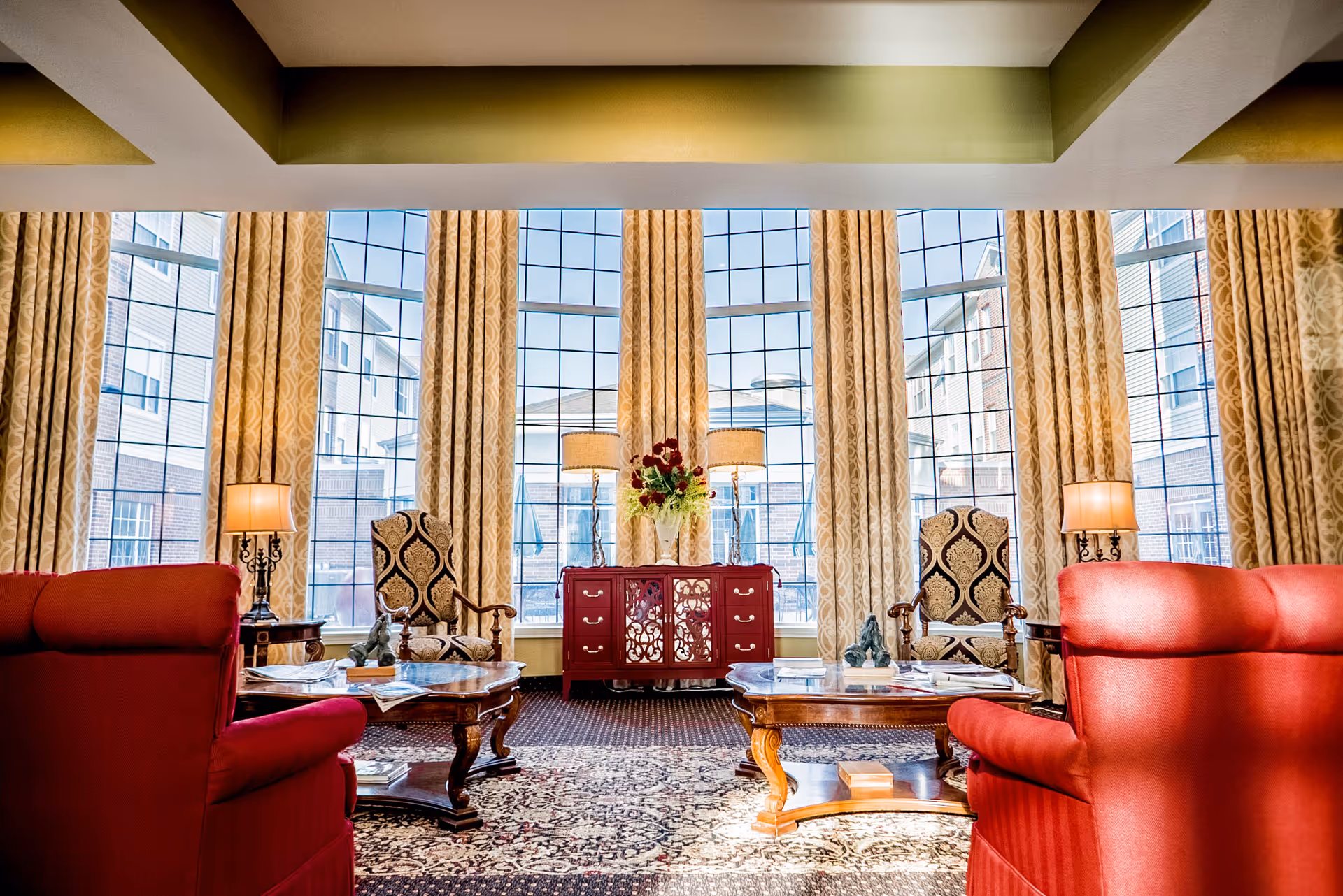 Bright and elegant living room area with large floor-to-ceiling windows covered by patterned curtains. The room features two red upholstered armchairs, two ornate wooden armchairs with patterned cushions, two wooden coffee tables with decorative items and magazines, a red sideboard with a floral arrangement and two lamps, and a patterned area rug on the floor.