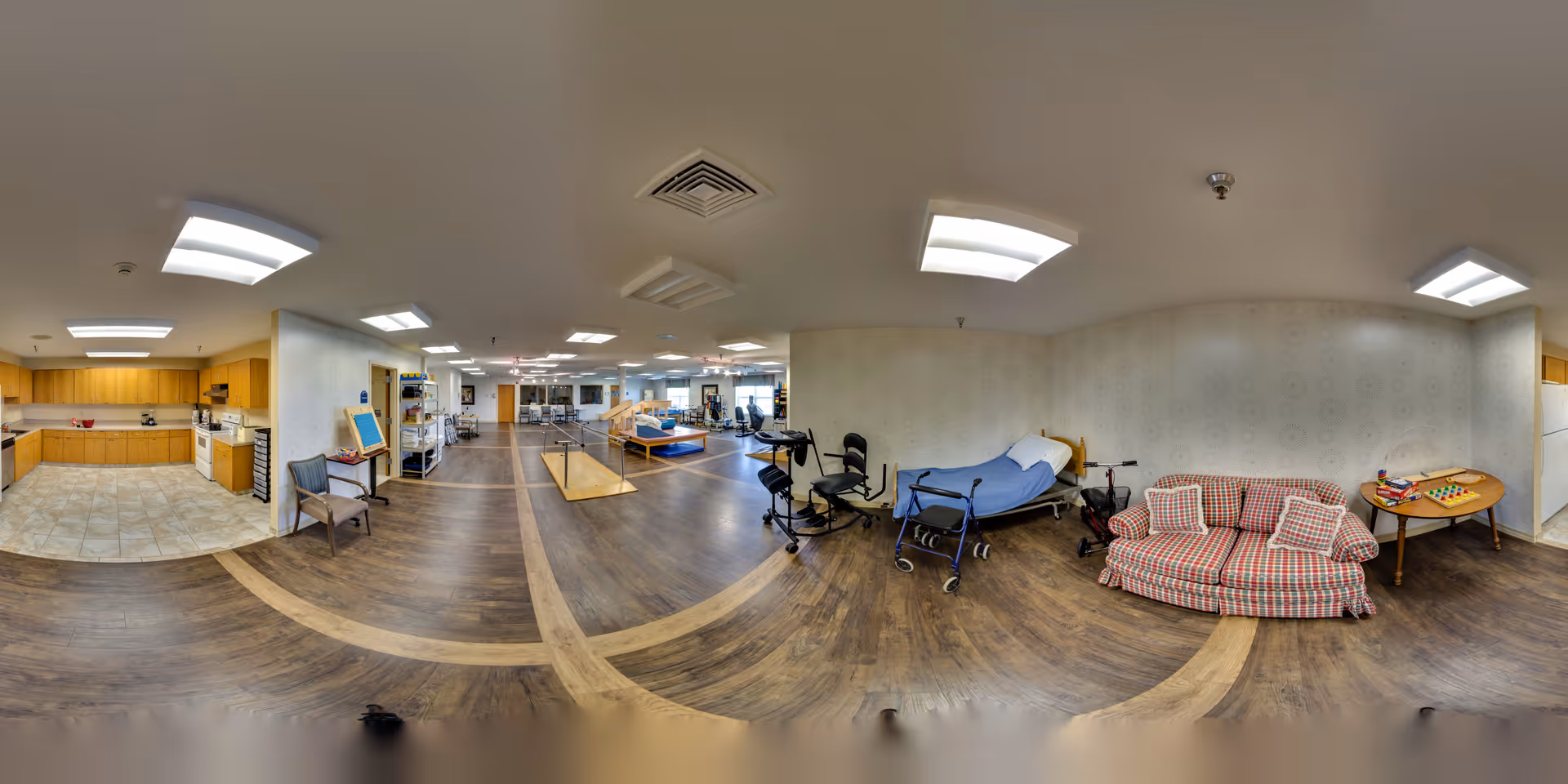 Wide-angle interior of a senior care facility showing a kitchenette, seating area with couch and table, a hospital bed and therapy equipment in a communal room.