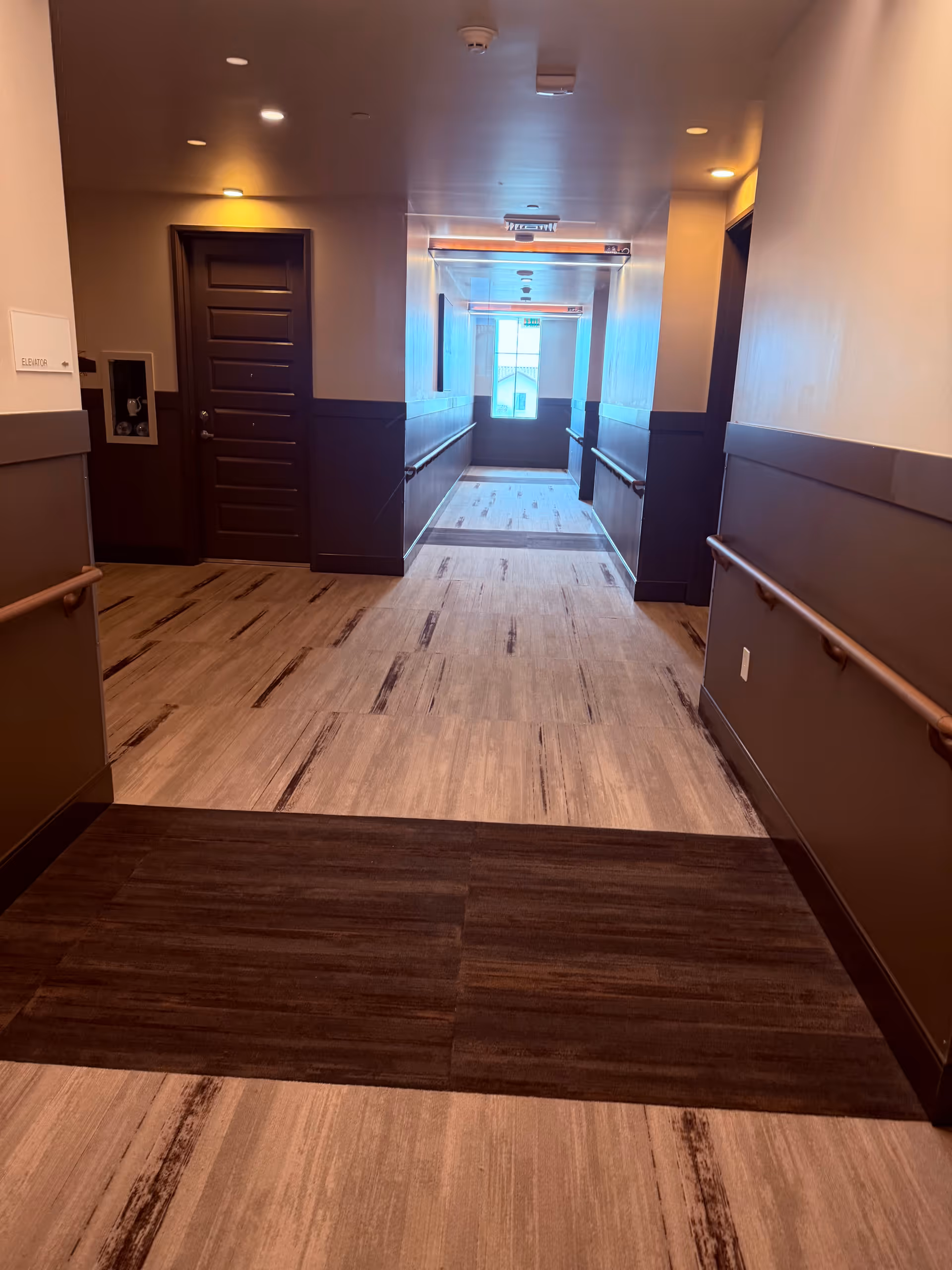Wide hallway inside a senior living facility with doors, wall rails, and warm overhead lighting.