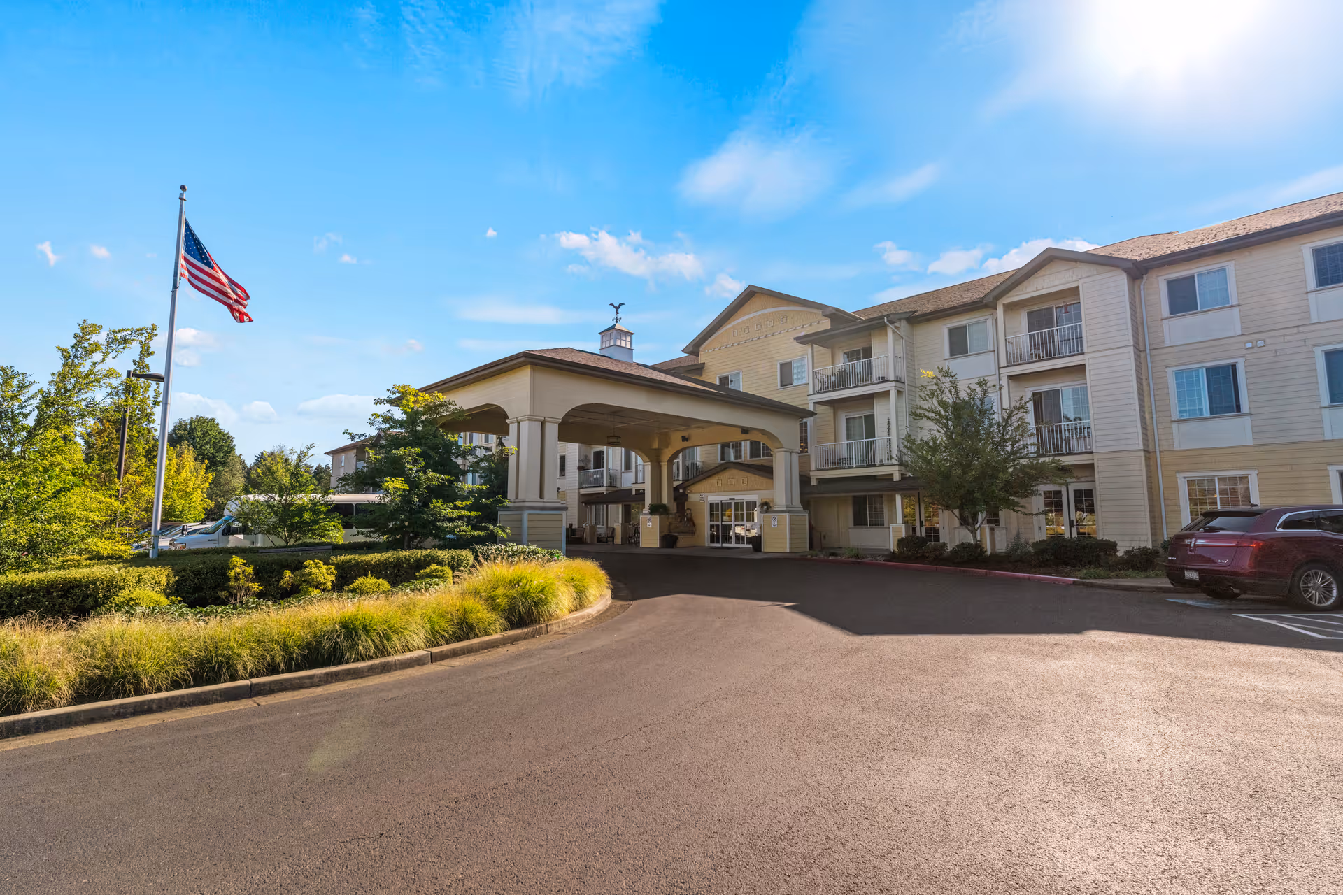 Exterior view of Volante Senior Living of Eugene building under a blue sky with some clouds. The building is three stories tall with balconies and large windows. There is a covered entrance driveway with columns, an American flag on a flagpole, landscaped greenery, and parked cars visible.
