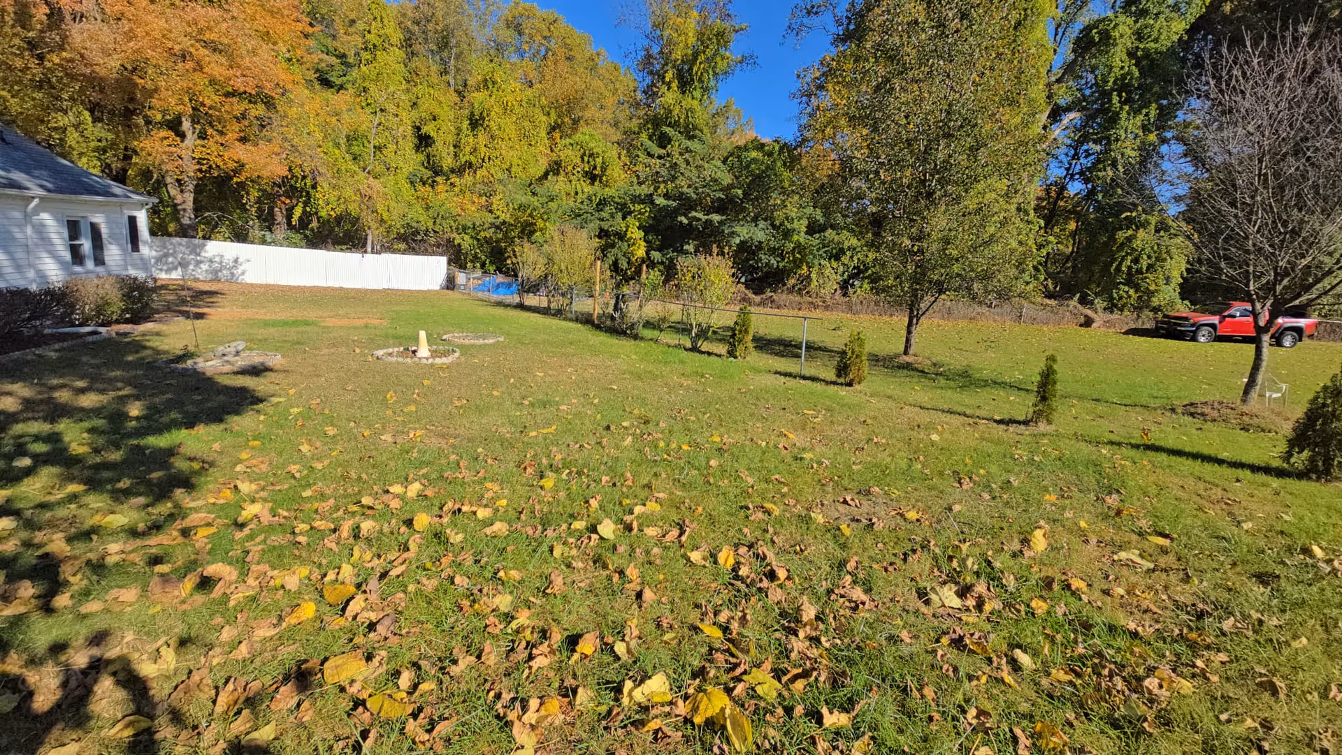 Wide grassy outdoor area with trees, fallen leaves, a white building, white fence, and trucks.