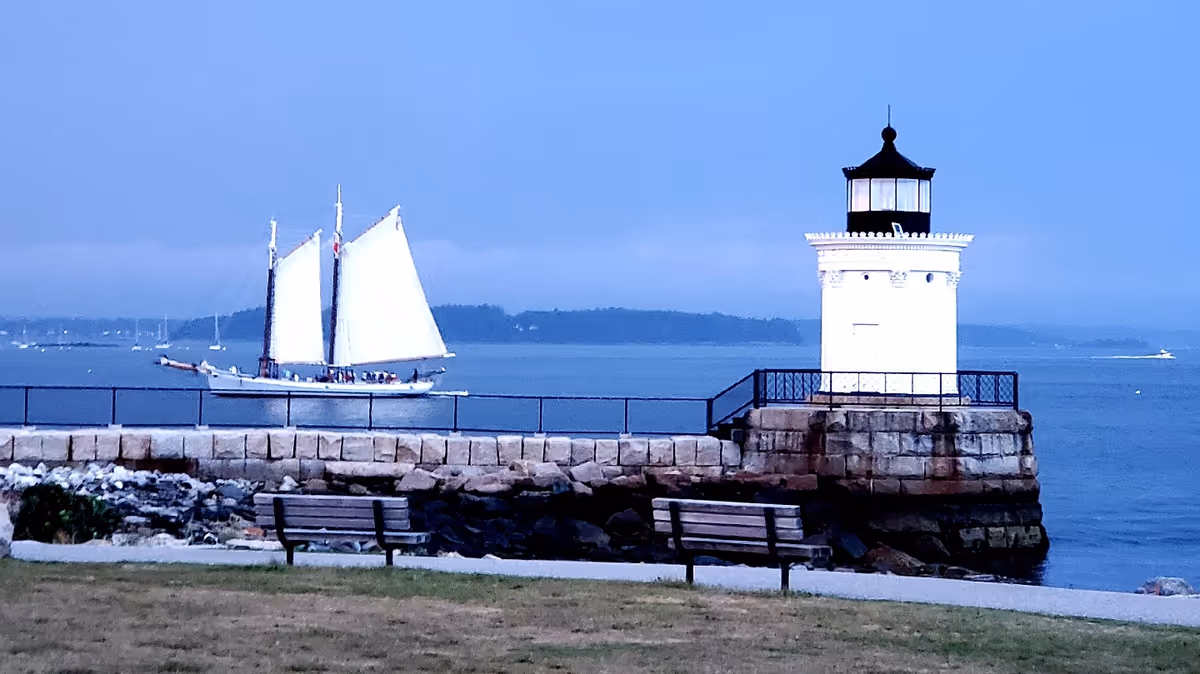 Lighthouse on a rocky pier with benches and a sailboat on calm water.