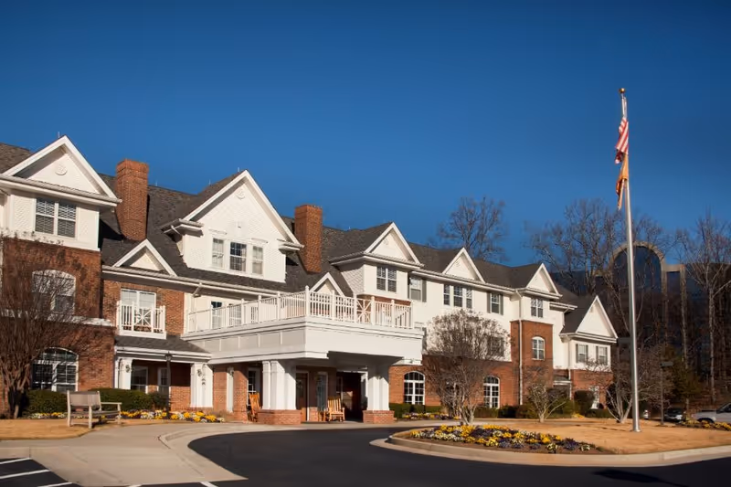 Front exterior of a multi-story brick-and-white senior living building with a covered entrance, flags, and landscaped driveway.