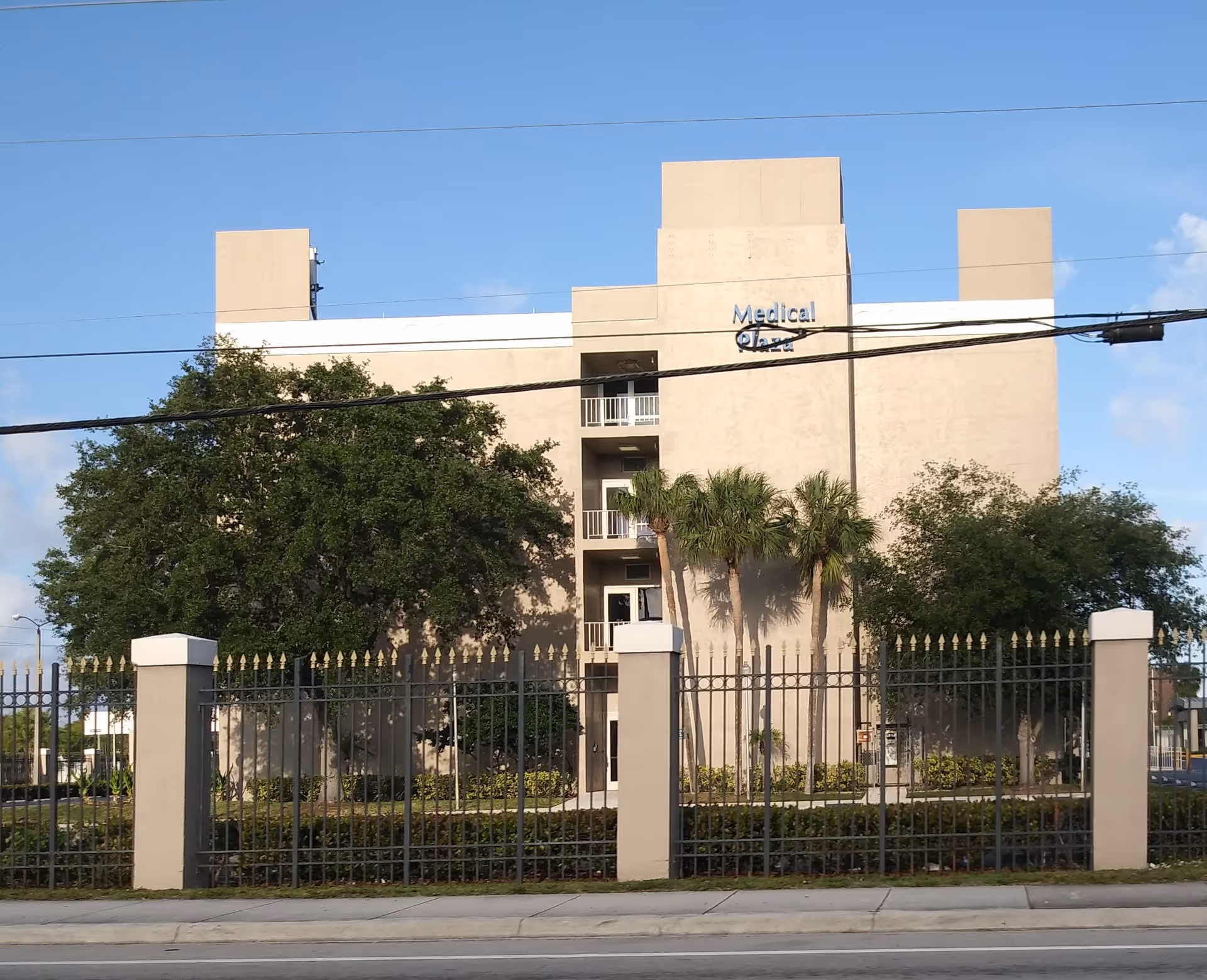Beige multi-story medical building seen behind a black metal fence and palm trees.