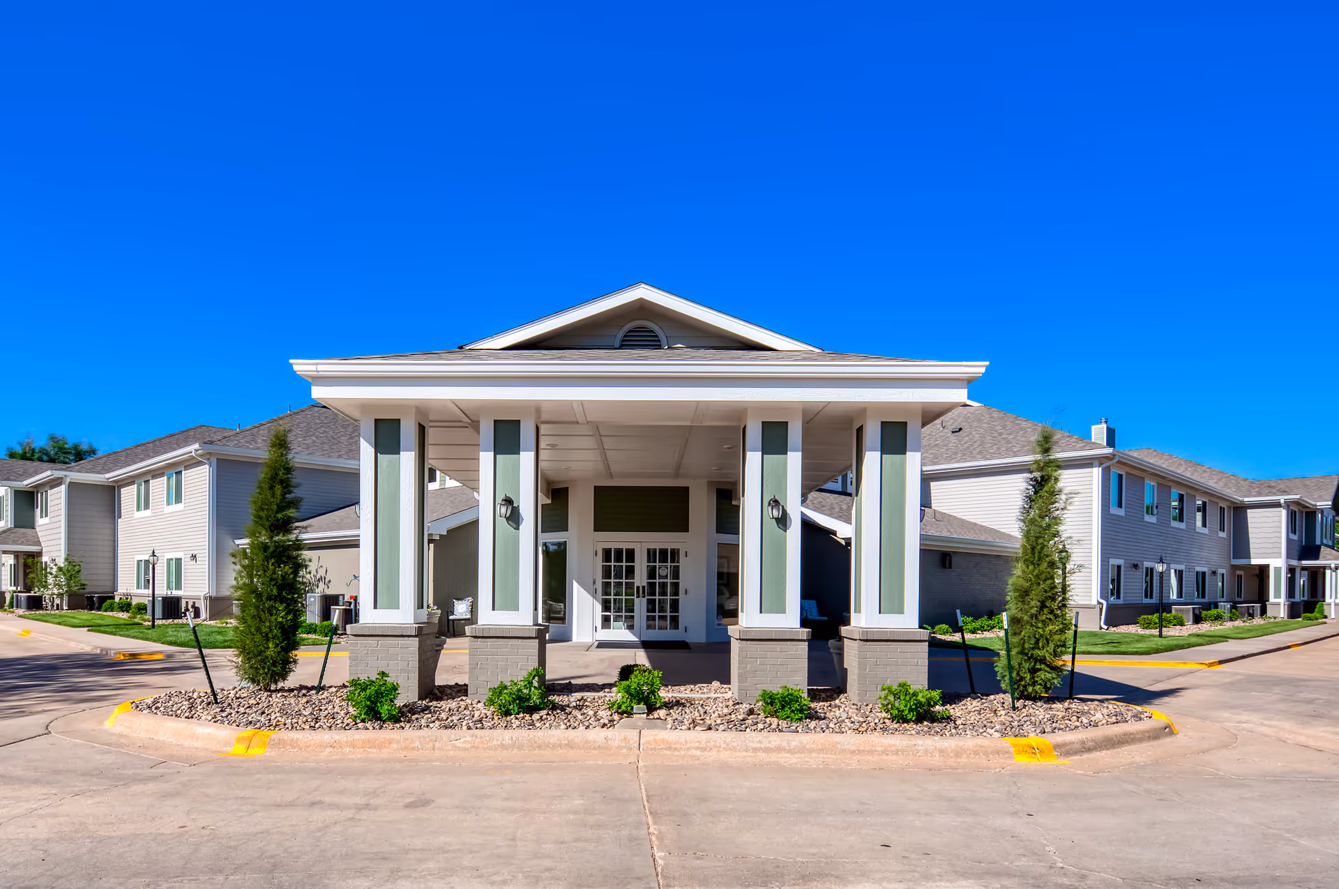 Front exterior view of Parkwood Village facility showing a covered entrance with pillars, landscaped area with small bushes and trees, and two-story buildings on either side under a clear blue sky.