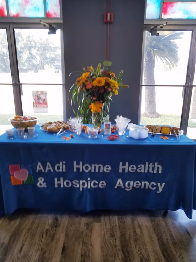 Indoor table with blue cloth reading AAdi Home Health & Hospice Agency, flower centerpiece, snacks.