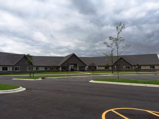 Exterior view of a single-story assisted living facility building with a large parking lot in front, small trees planted along the sidewalk, and a cloudy sky overhead.