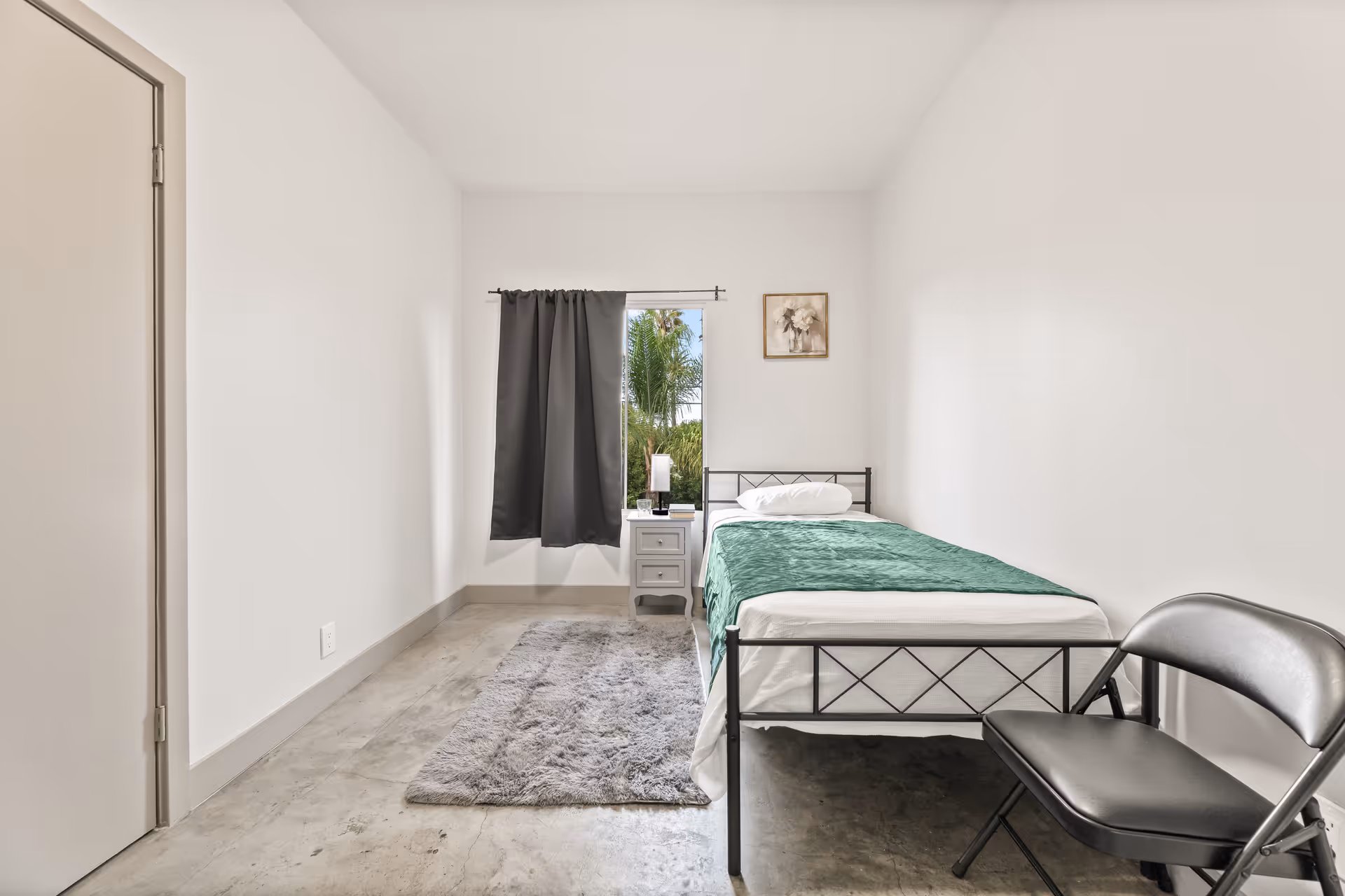 Minimal bedroom with a single bed, nightstand, window, rug, and folding chair.