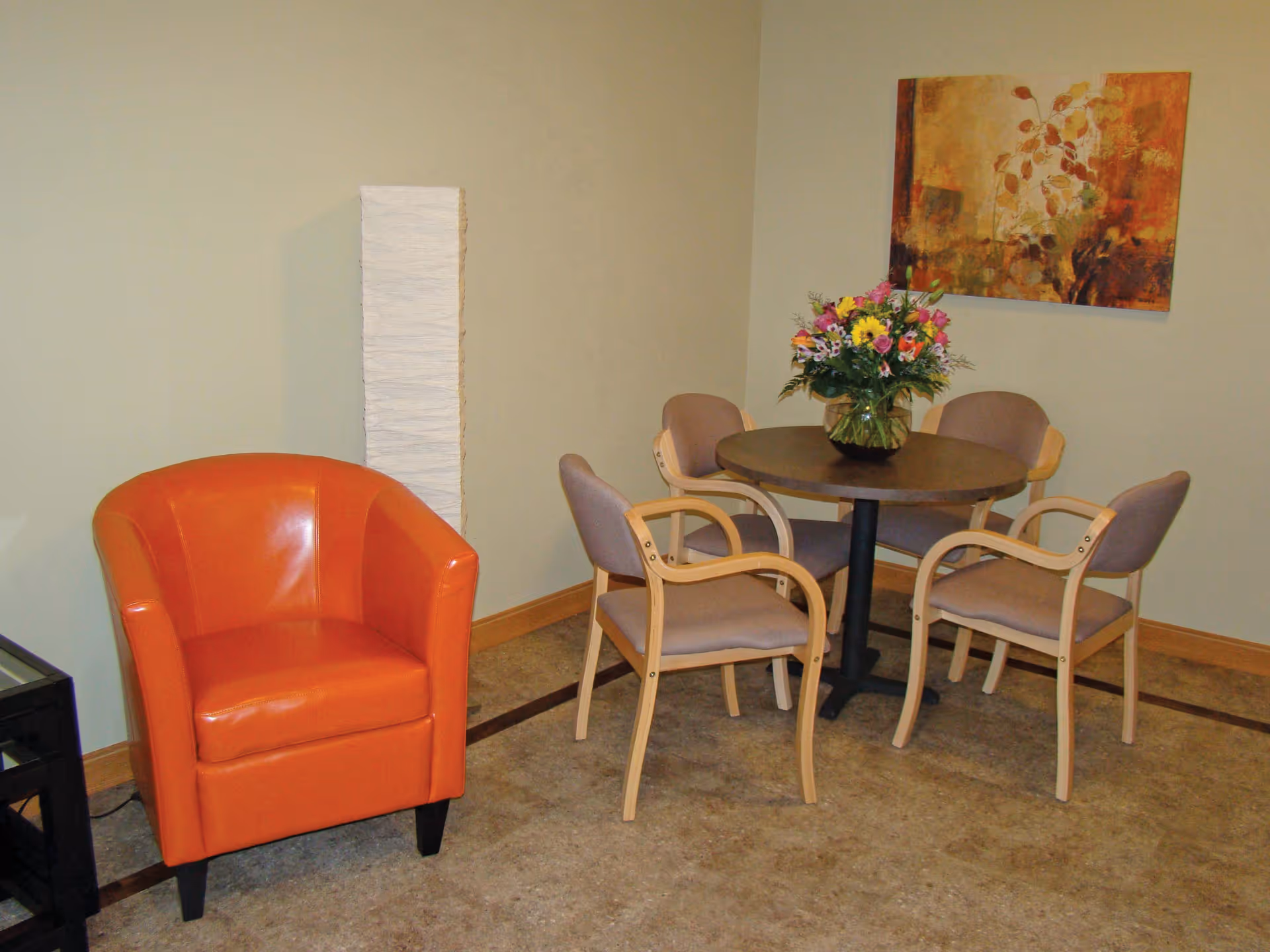 A small seating area with a round wooden table surrounded by four beige cushioned chairs with wooden arms. A vase with a colorful bouquet of flowers is placed on the table. To the left of the table is an orange leather armchair. A tall, narrow white floor lamp stands against the wall behind the armchair. A framed abstract painting with warm tones hangs on the wall above the table.