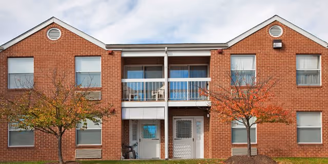Exterior view of a two-story red brick building with white trim and a central balcony. There are two small trees with autumn-colored leaves in front of the building, and the sky is partly cloudy.