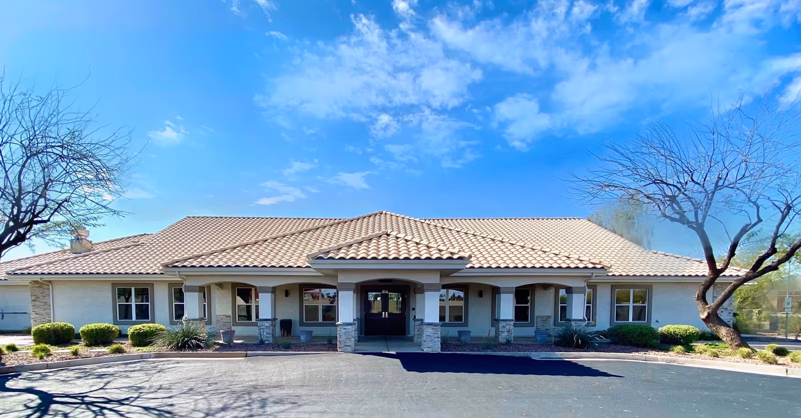 Front exterior view of a single-story building with a tiled roof, multiple windows, and a covered entrance supported by stone pillars. The building is surrounded by landscaped bushes and trees under a blue sky with scattered clouds.