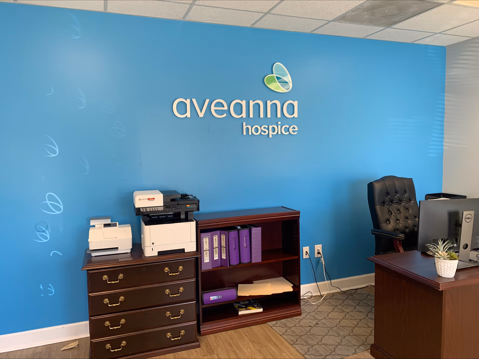 Blue wall reception area with Aveanna Hospice logo, desk, printer, and filing cabinet.