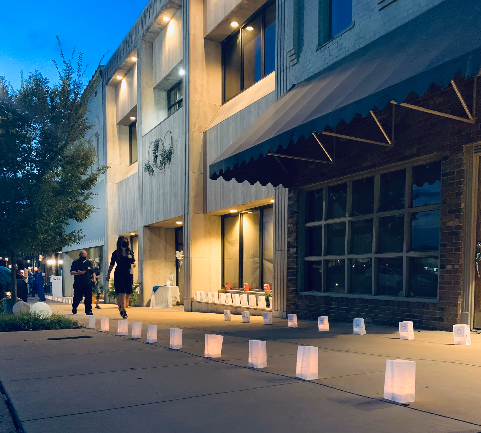 Dusk exterior of a modern building with an awning, lantern-lined sidewalk, and people walking.