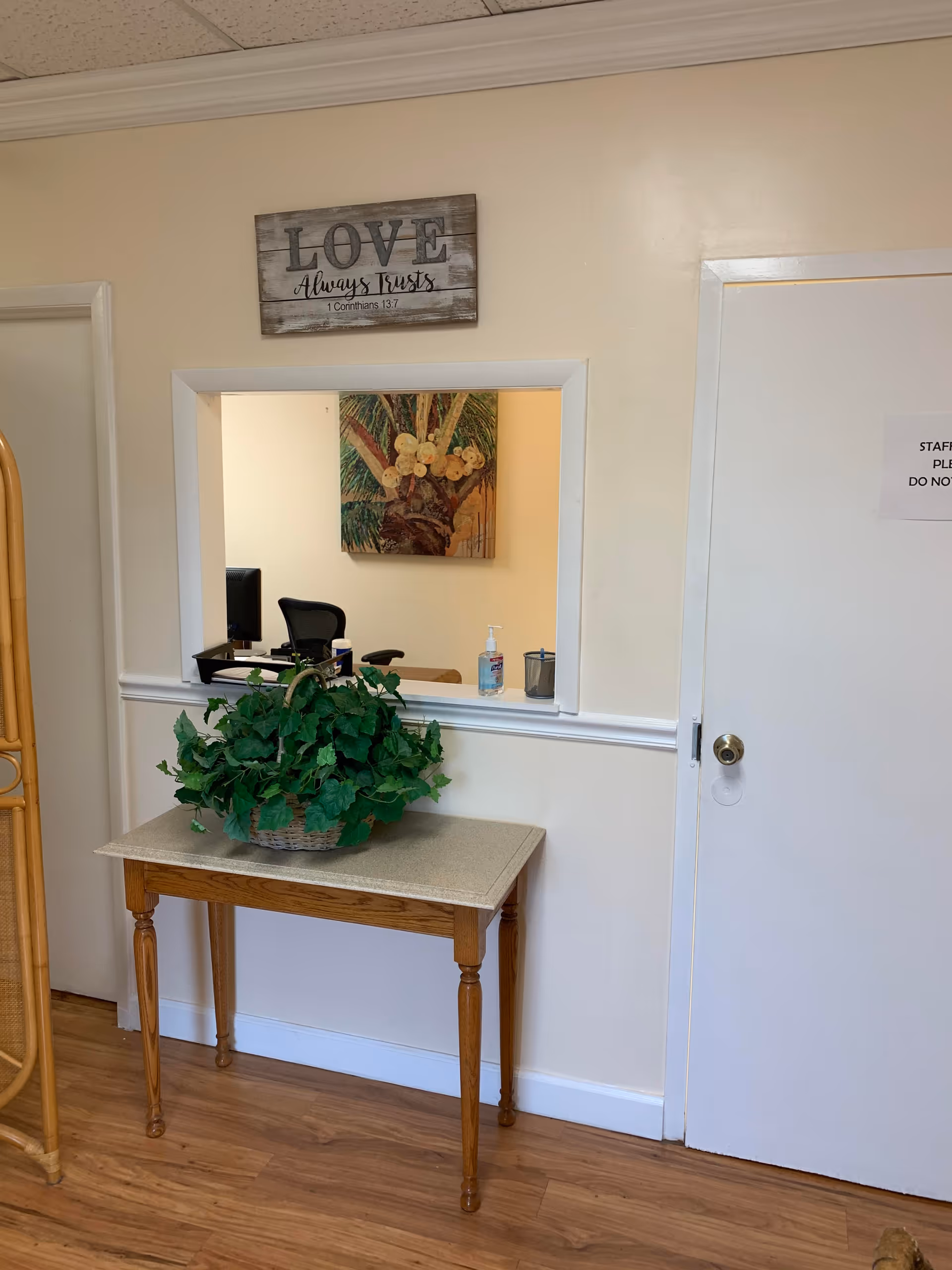 Indoor reception area with a pass-through window, wooden table, and potted plant.