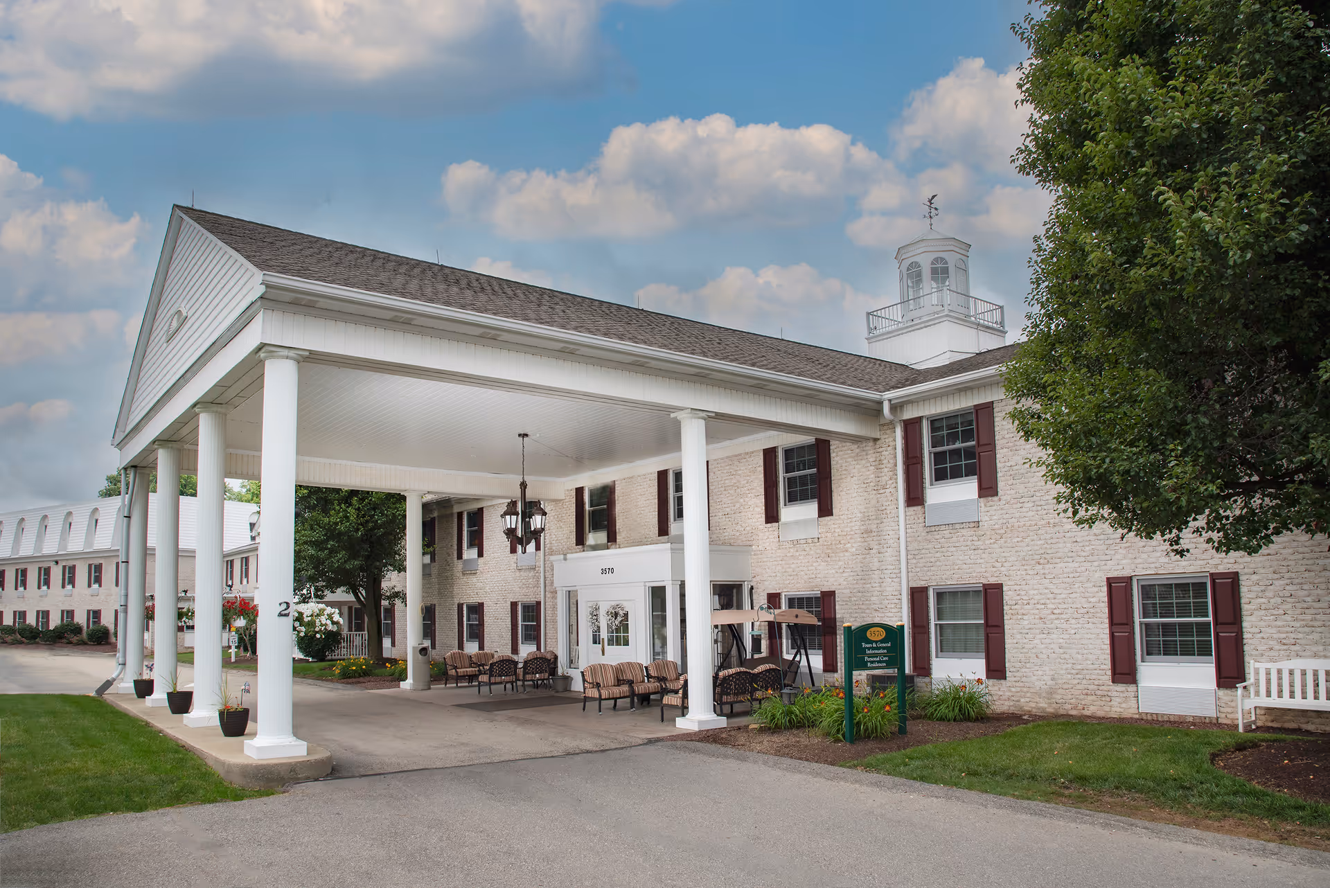 Exterior view of a senior living facility building with white brick walls, maroon shutters, and a covered entrance supported by white columns. There are benches and chairs under the covered area, a green lawn, and a tree on the right side. The sky is partly cloudy.
