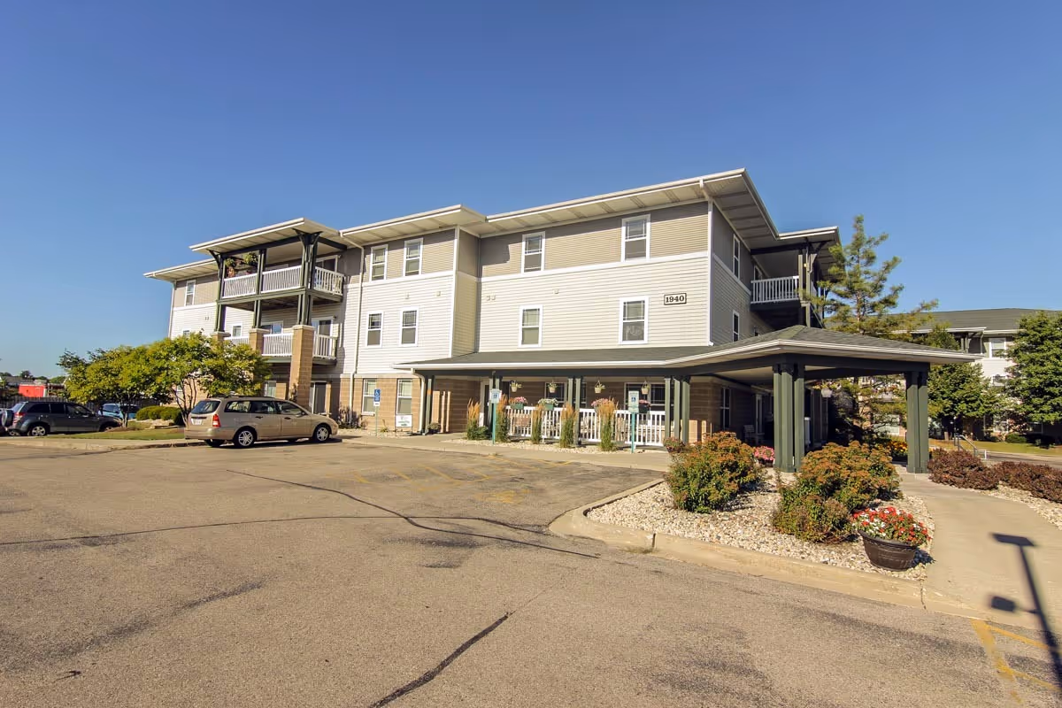Exterior view of a three-story senior apartment building with balconies, a covered entrance, parked cars, and landscaped bushes and flowers under a clear blue sky.