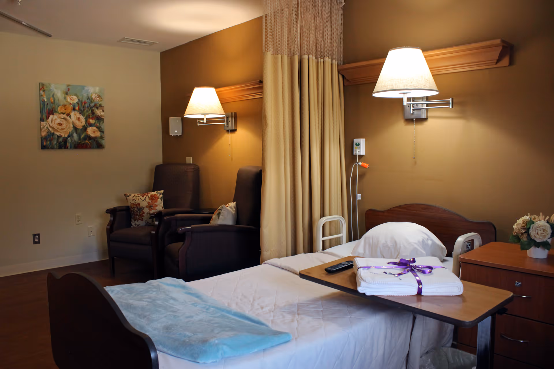 A cozy senior living facility bedroom with a single bed covered in white bedding and a light blue blanket. A wooden overbed table holds folded towels tied with a purple ribbon and a remote control. Two dark armchairs with floral cushions are positioned near a wall with a floral painting. The room is softly lit by two wall-mounted lamps, and a beige privacy curtain hangs between the bed and chairs. A small wooden nightstand with a flower arrangement is beside the bed.