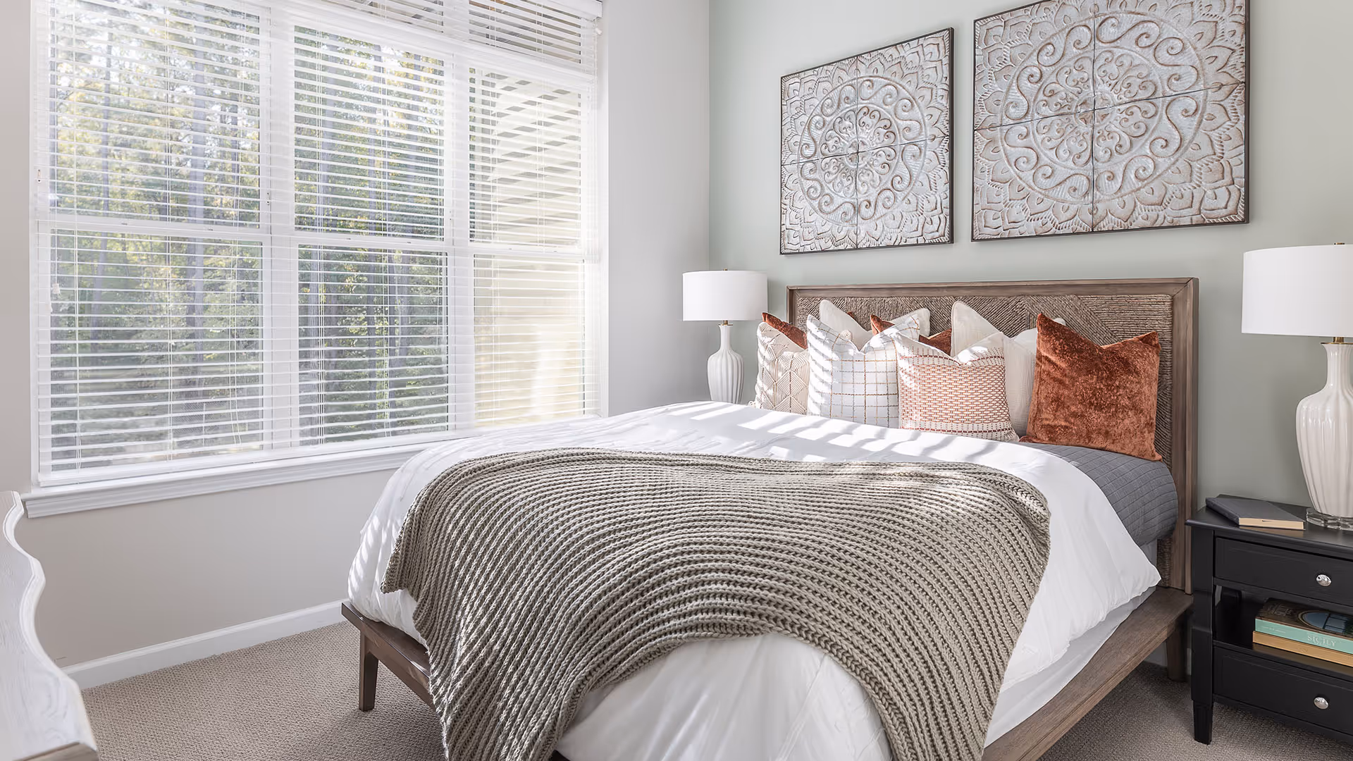 A bright bedroom with a large window covered by white blinds letting in natural light. The bed has a wooden frame and is neatly made with white bedding, a textured gray blanket, and multiple decorative pillows in shades of white, gray, and rust. On either side of the bed are matching white lamps on black nightstands. Above the bed are two decorative carved wooden wall panels.