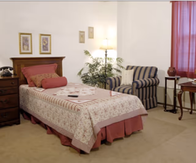 Single-occupancy bedroom with a neatly made twin bed, wooden nightstand, striped armchair, floor lamp and potted plant.