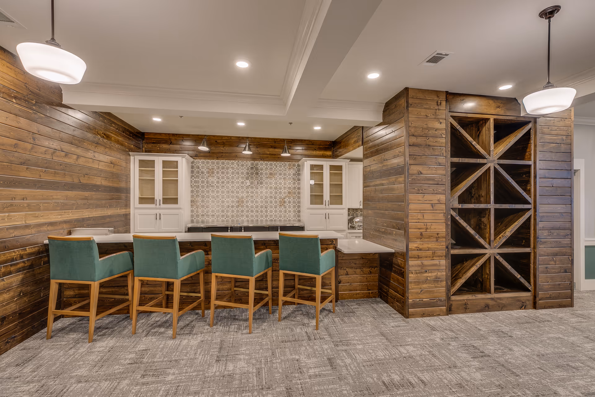 Interior view of a modern kitchen area with wooden walls and cabinetry. There is a countertop with four green cushioned bar stools, pendant lights hanging from the ceiling, and a large wooden wine rack on the right side. The floor is carpeted with a patterned design.