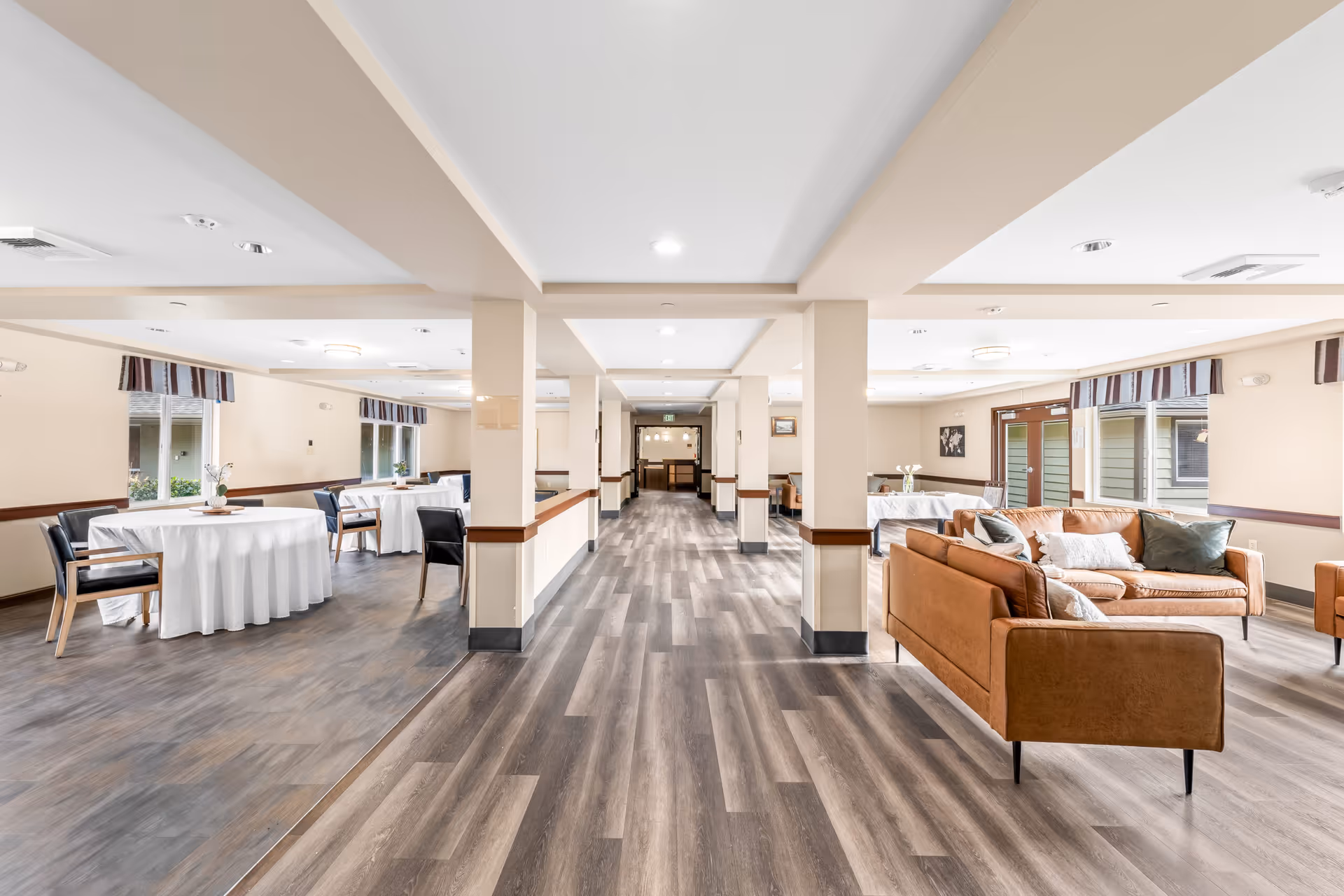 Bright indoor senior living common area with brown sofas, round tables, and a long central hallway.