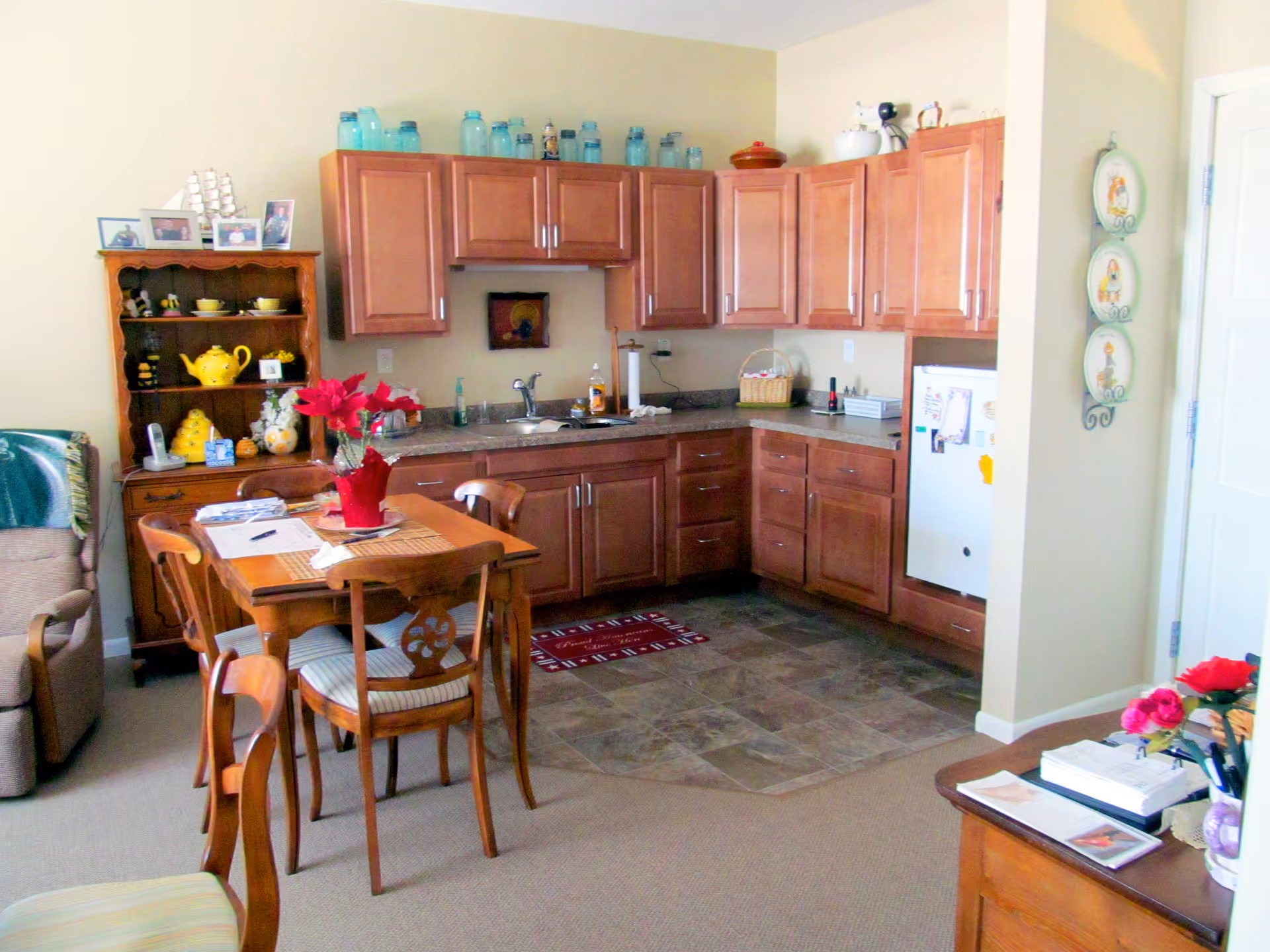 A cozy kitchen and dining area with wooden cabinets, a small white refrigerator, a wooden dining table with four chairs, and various decorative items including a red poinsettia centerpiece on the table and blue jars on top of the cabinets.