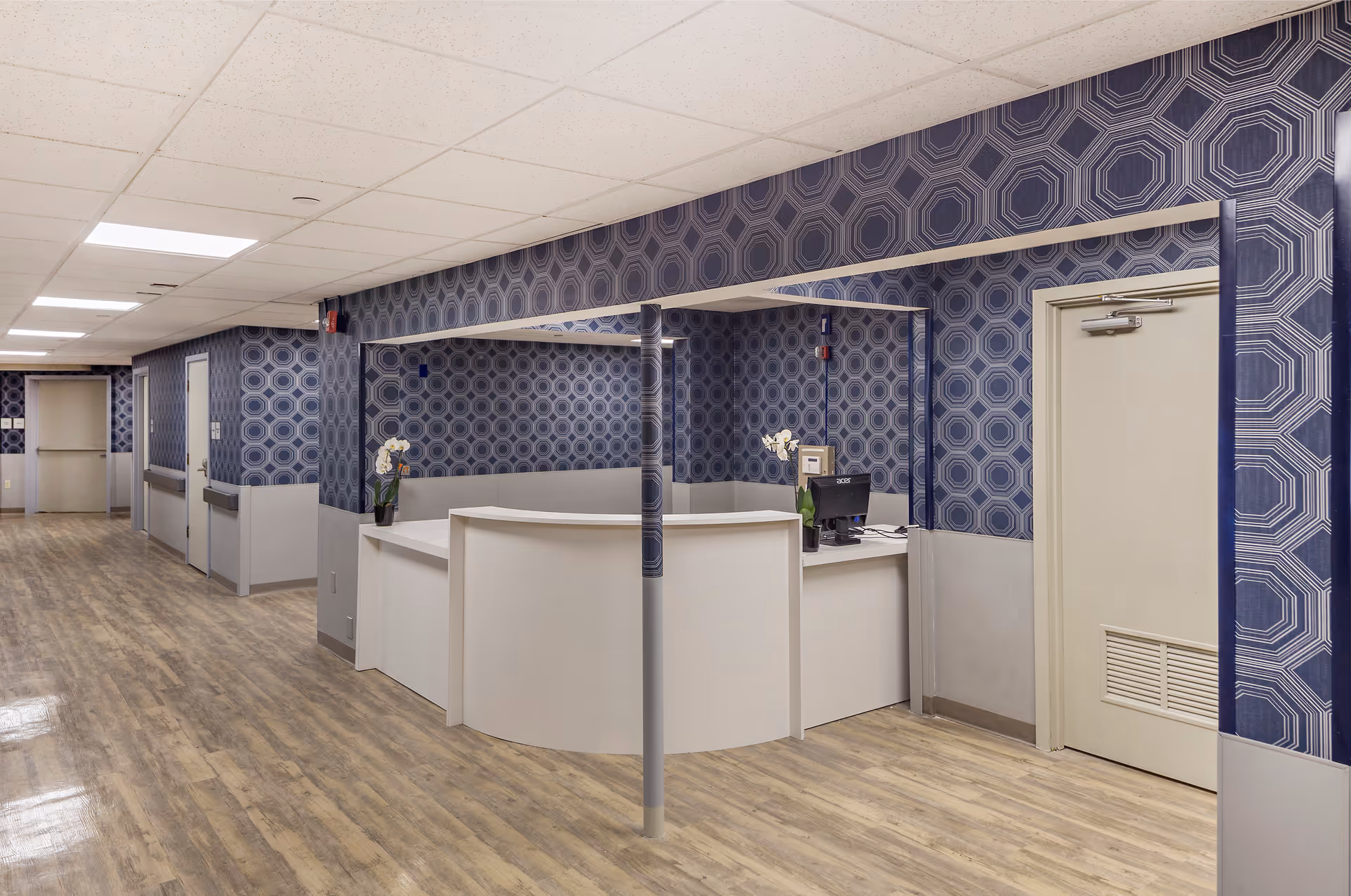 Interior view of a senior living facility hallway with a reception desk area. The walls are decorated with blue geometric patterned wallpaper, and the floor has a light wood finish. The reception desk is white with a computer and a small plant on it. There are several doors along the hallway and ceiling lights illuminating the space.
