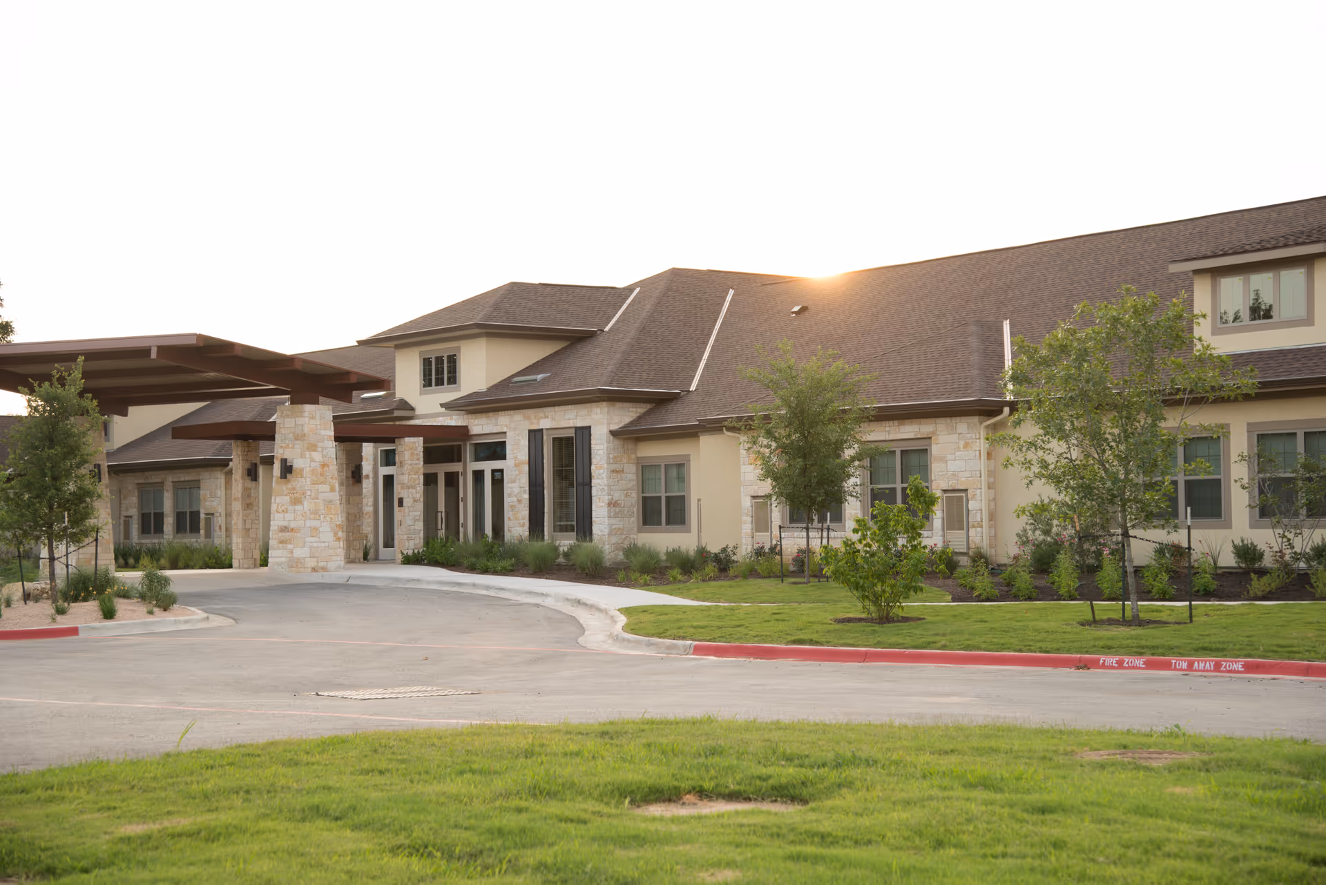 Front exterior of a single-story memory care facility with stone and stucco facade, covered entry canopy, curved driveway, and landscaped lawn.