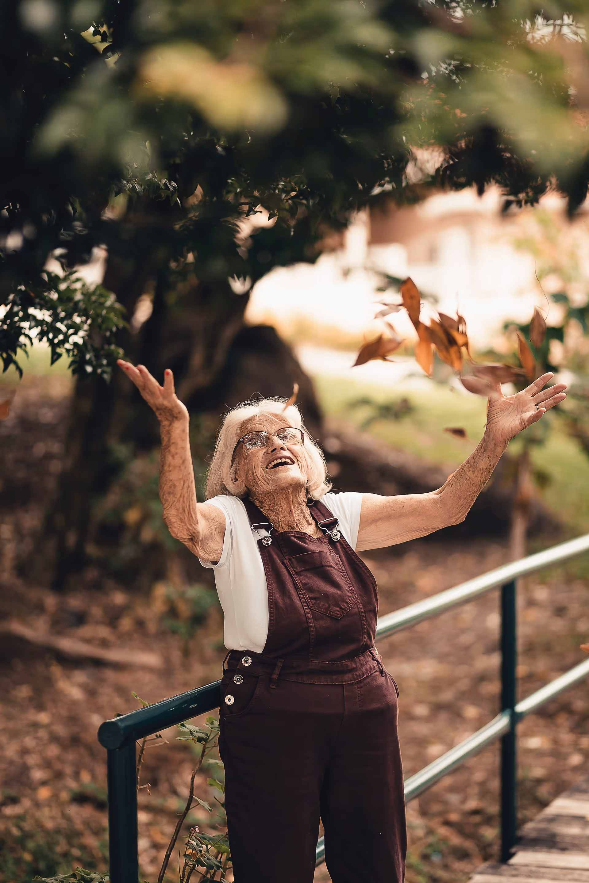 Smiling elderly woman outdoors, arms raised, tossing autumn leaves near a railing.