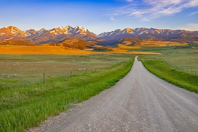 Gravel road winds through green fields toward snow-capped mountains under blue sky.
