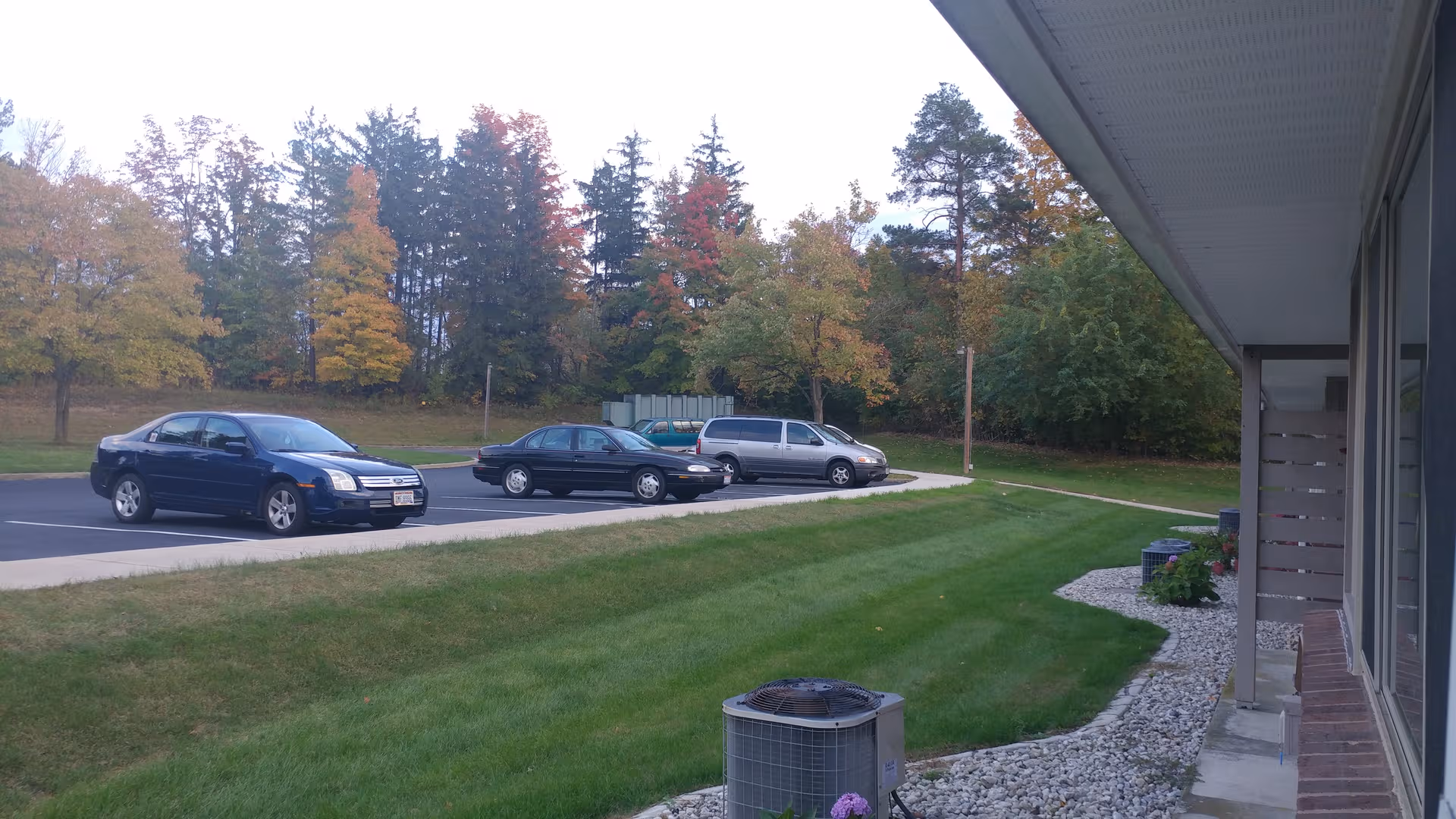 Senior living facility exterior with parked cars and autumn trees by a manicured lawn.