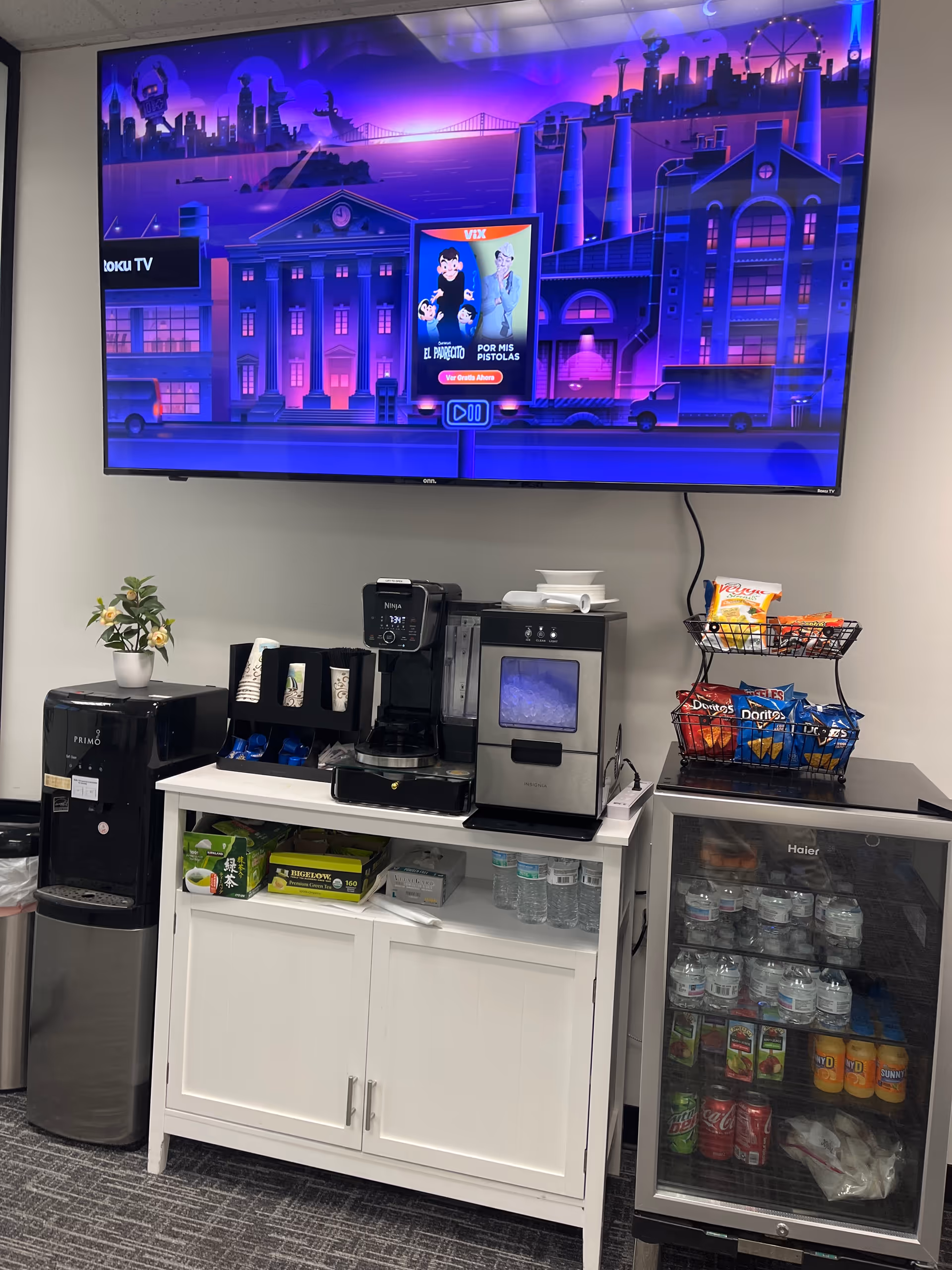 A wall-mounted TV above a kitchenette counter with coffee machine, snacks, and beverages.