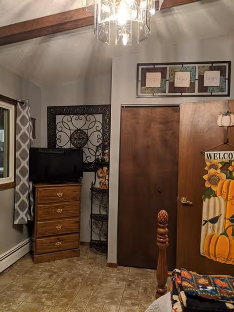 A cozy bedroom corner with a wooden dresser holding a small TV, a decorative metal wall hanging above it, and a three-tiered metal shelf with small decorations. There is a window with patterned curtains on the left, a wooden closet door in the center, and a partially open wooden door on the right with a fall-themed welcome sign featuring pumpkins. Above the closet door is a framed artwork with the words 'Faith', 'Love', and 'Hope'. The floor is tiled, and part of a wooden bedpost and a colorful blanket are visible in the foreground.