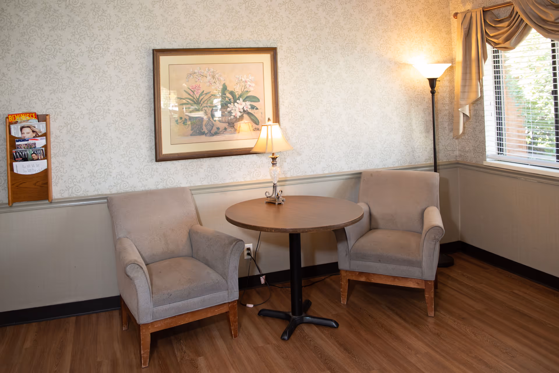 Cozy indoor seating area with two beige armchairs, a round table, lamp, and patterned wallpaper.